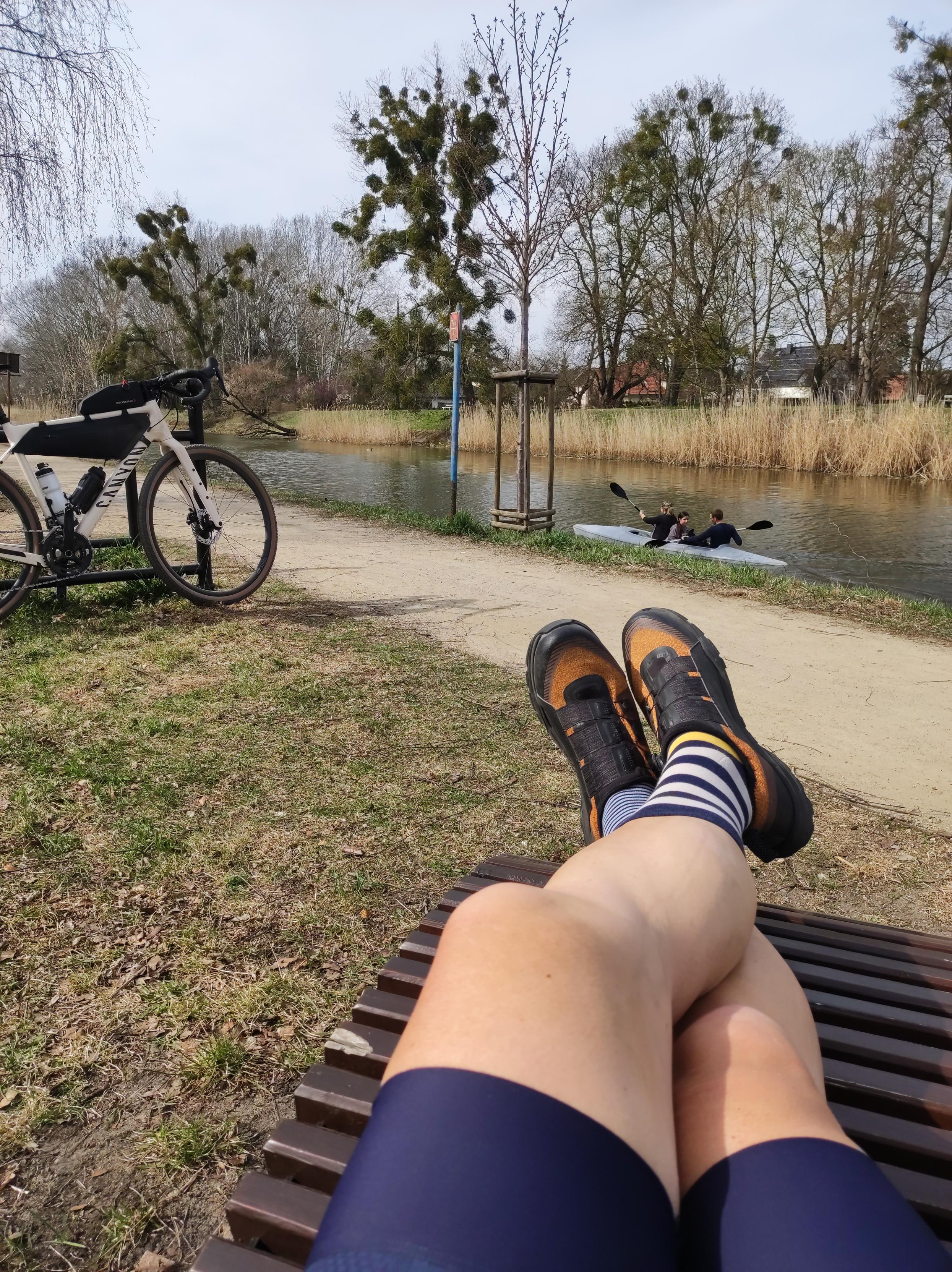 Słoneczny dzień. Moje nogi w kolarskich butach i spodenkach wyciągnięte na drewnianym leżały nad brzegiem kanału. Przy stojaku po lewej stoi mój rower,  a kanałem płynie kajak z trzema osobami.