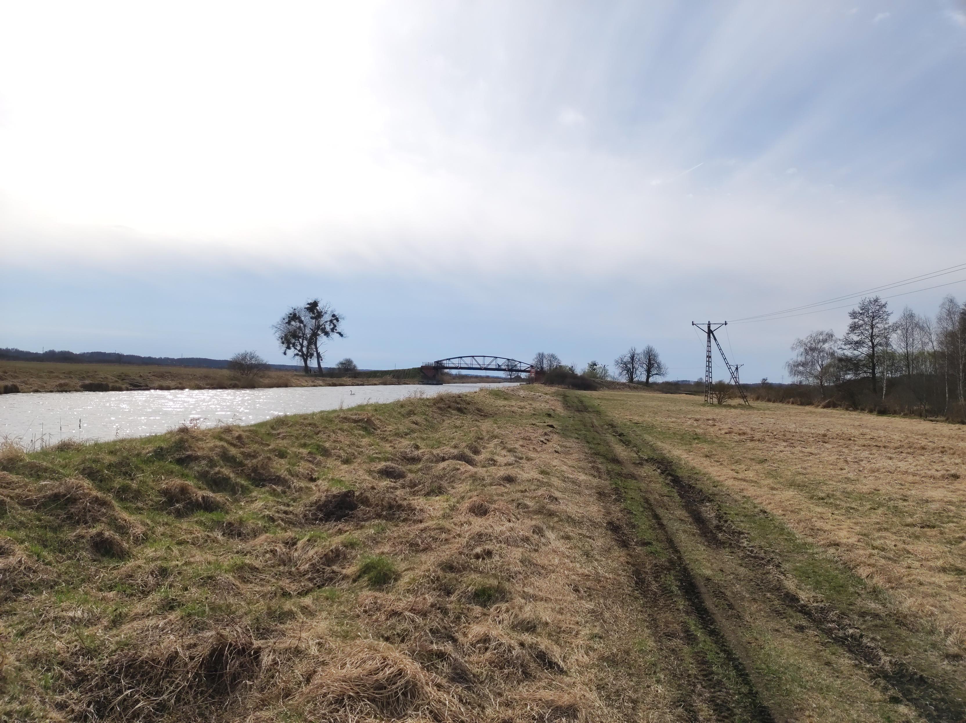 Słabo widoczna droga przez łąki wzdłuż widocznego po lewej kanału. W oddali most nad kanałem.