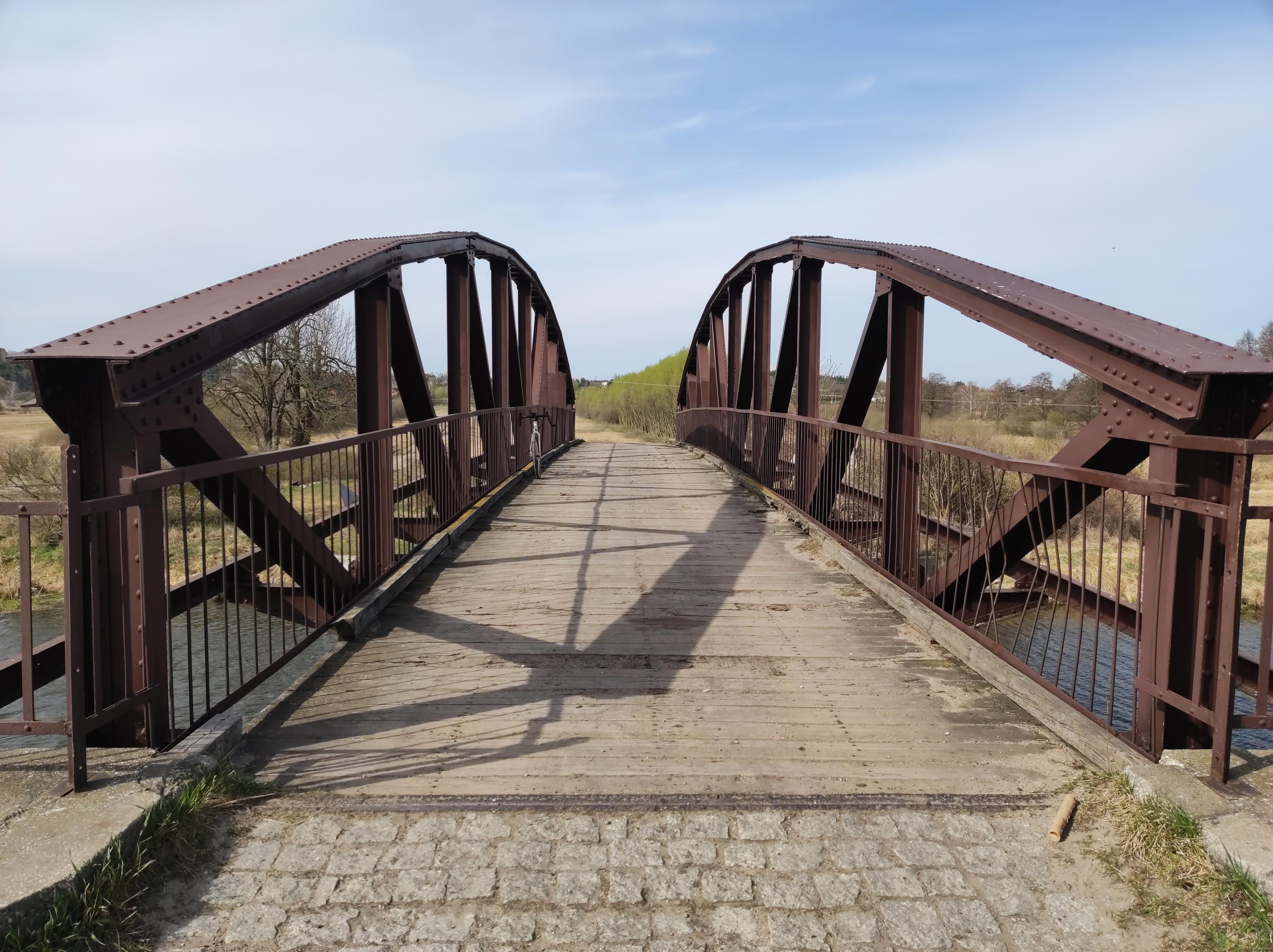 Wyłożony drewnianymi deskami metalowy most, do którego prowadzi krótki pasek bruku.