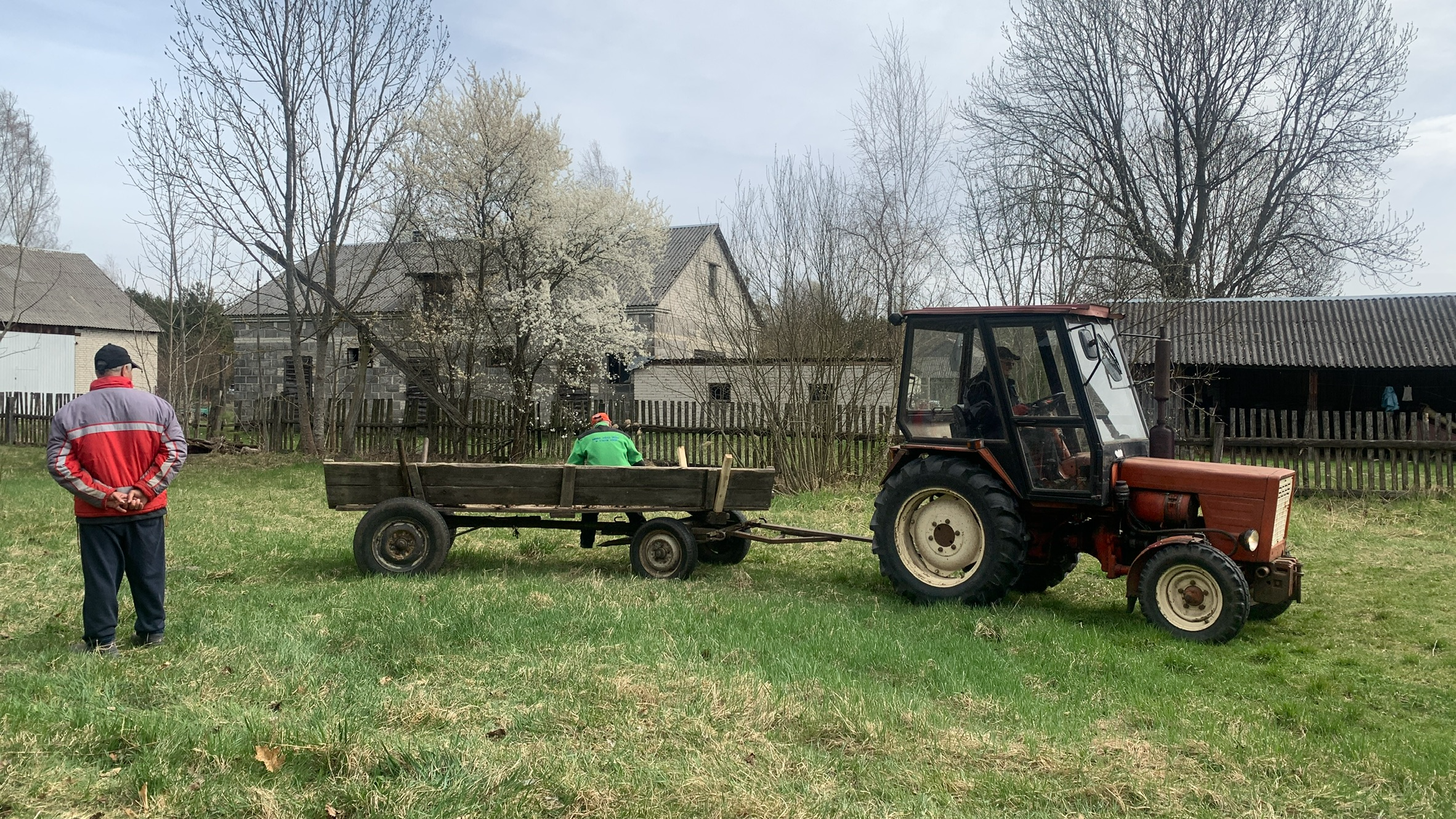 Pusta furmanka za traktorem, siedzi na niej bokiem mężczyzna. W tle zabudowania gospodarcze