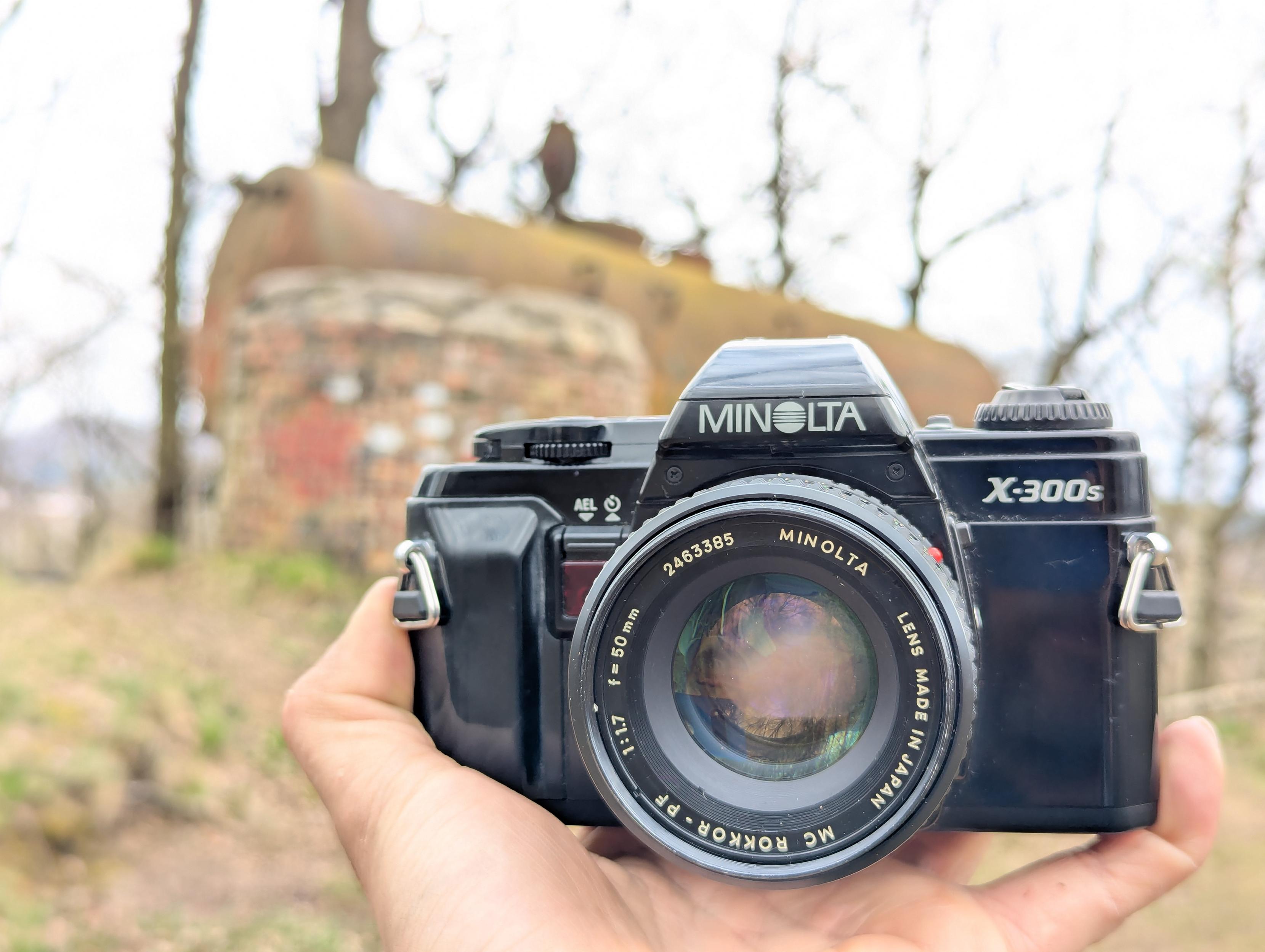 zdjęcie wyciągniętej dłoni trzymającej minoltę x-300s z obiektywem 50mm; w tle widać kontur stalowego zbiornika niewiodomego przeznaczenia