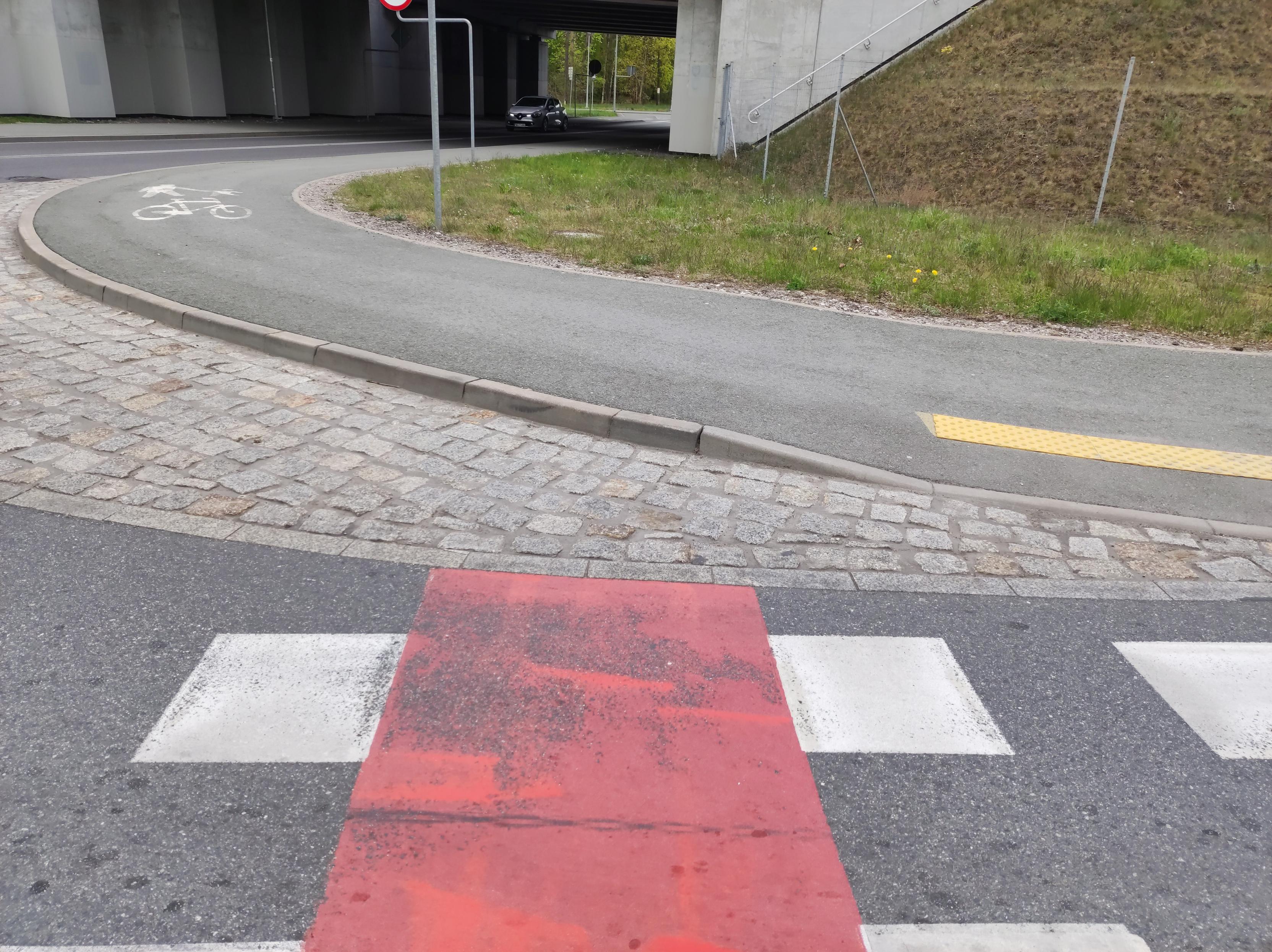 Przejazd dla rowerów obok przejścia dla pieszych, prowadzący wprost na wyłożony brukiem łuk na skrzyżowaniu i wysoki krawężnik.