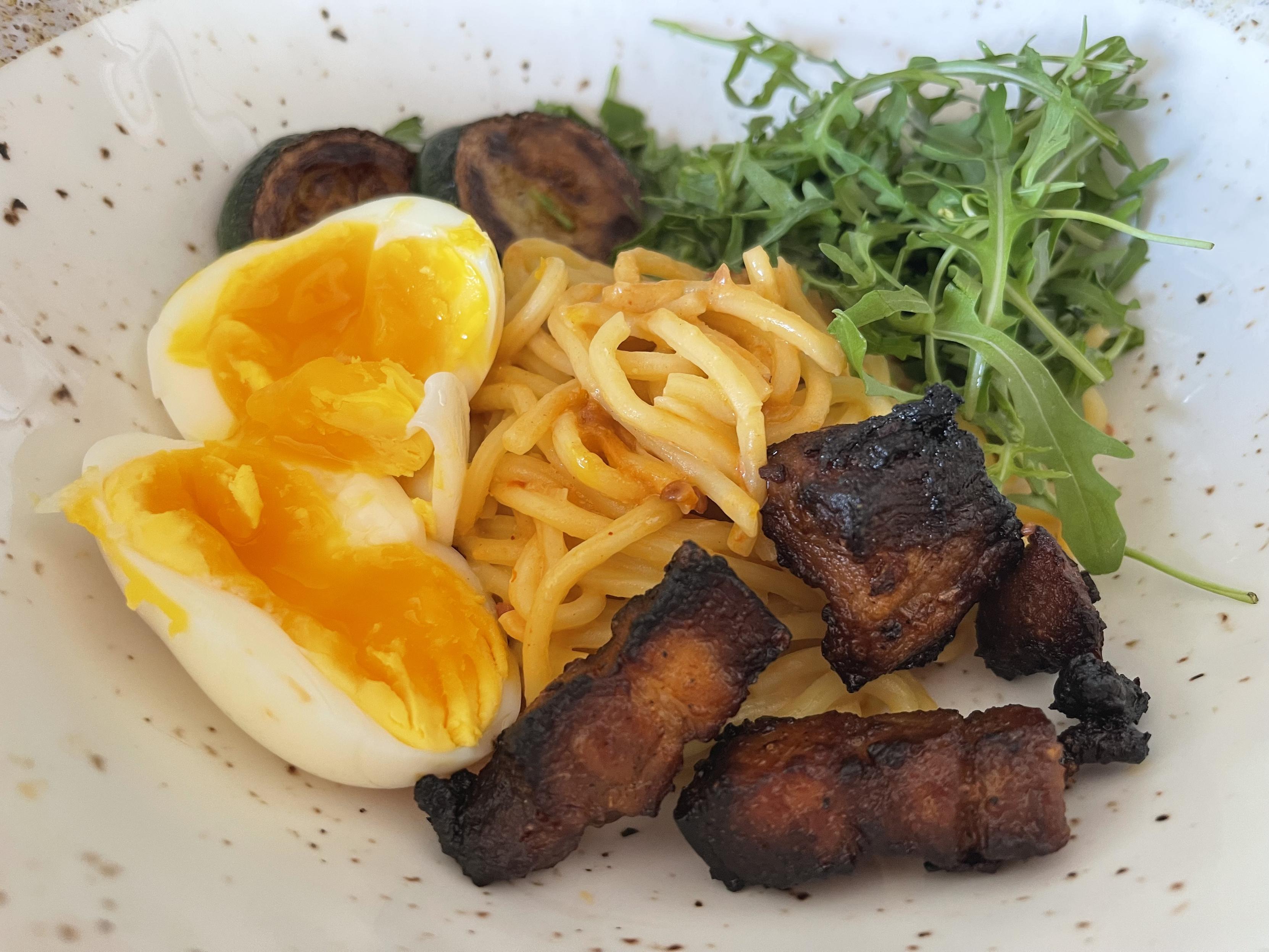 Na jasnym, nakrapianym talerzu znajduje się porcja makaronu ramen w pomarańczowym sosie miso. Obok leży przekrojone jajko na półmiękko z intensywnie żółtym, lekko płynnym żółtkiem, kawałki mocno przypieczonego chrupiącego boczku, plasterki podsmażonej cukinii oraz garść świeżej rukoli.