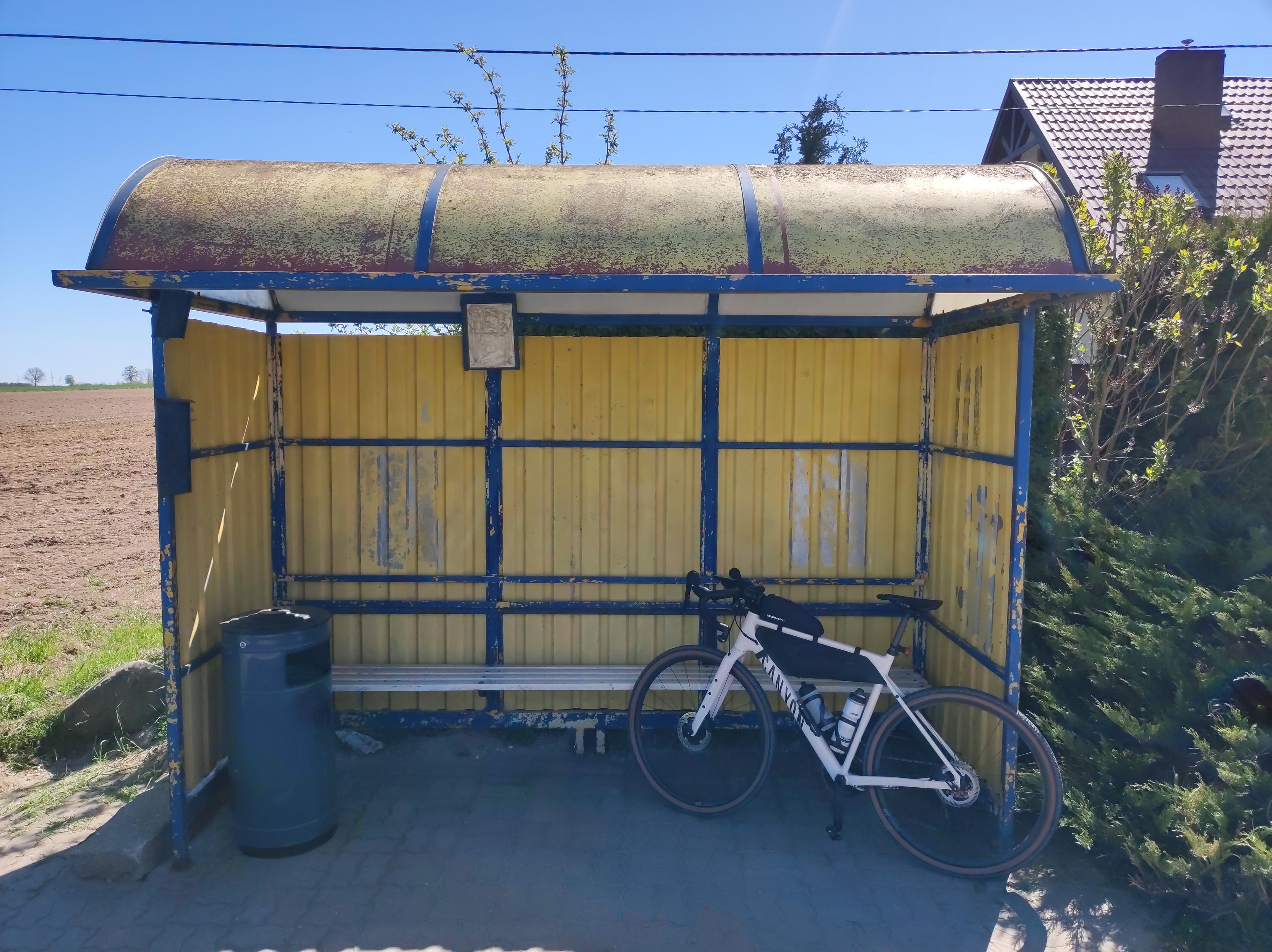 Żółta wiata przystankowa, o którą opiera się jasny rower szutrowy Canyon. W tle po prawej widać kawałek dachu wystający z krzaków, a po lewej puste pole.