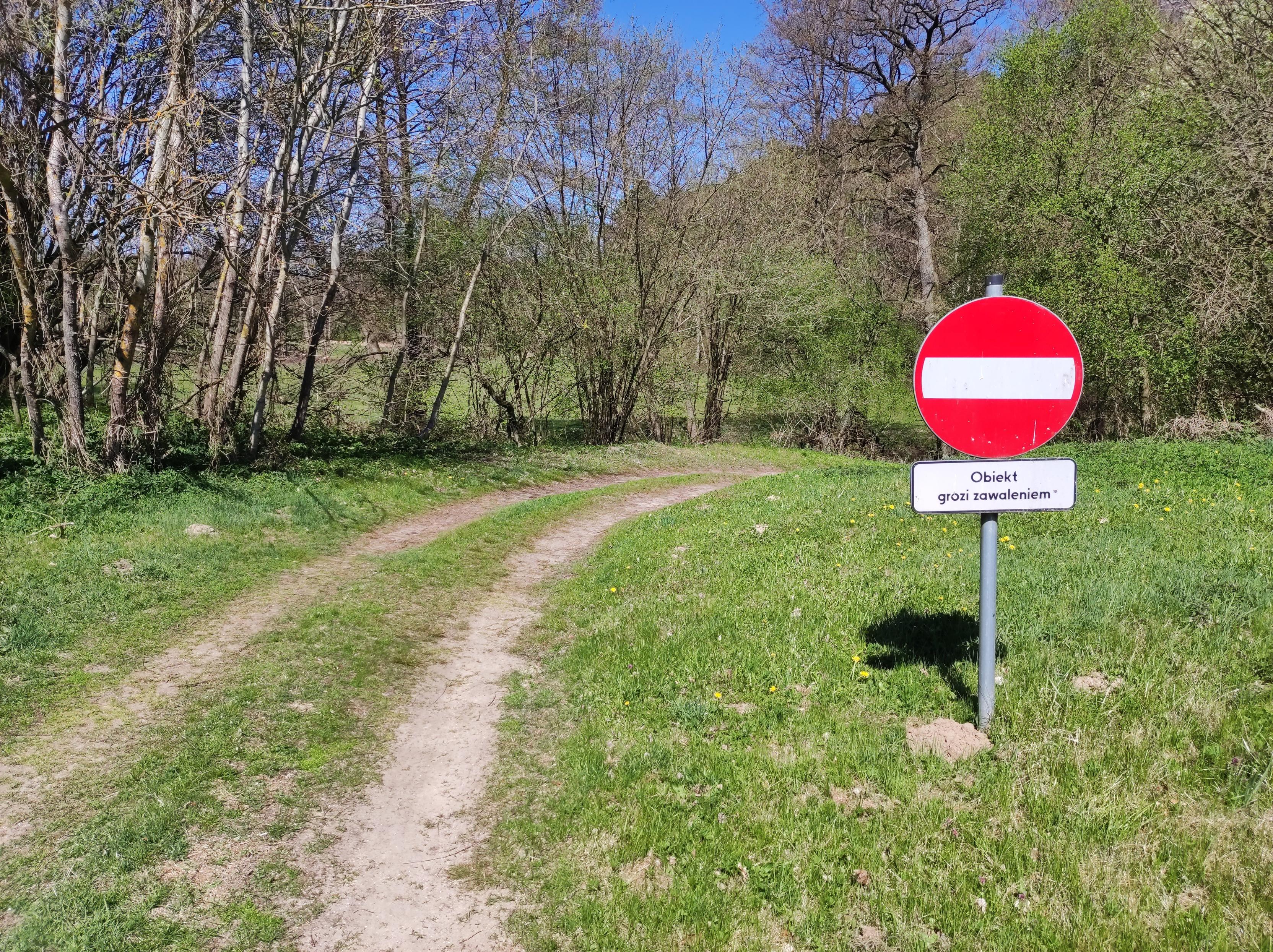 Droga przez łąkę skręcająca w prawo za znakiem zakazu wjazdu z tabliczką "Obiekt grozi zawaleniem"