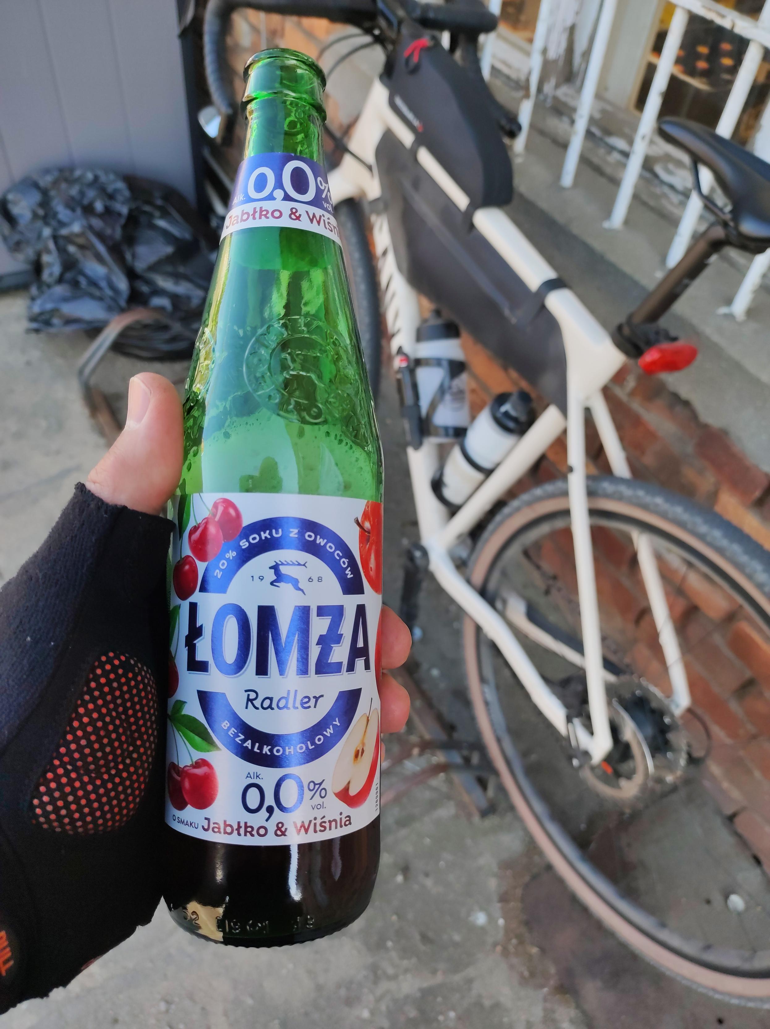 Ręką w kolarskiej rękawiczce trzyma butelkę radlera 0% z Łomży o smaku jabłko i wiśnia. W tyle oparty o ścianę sklepu stoi mój rower
