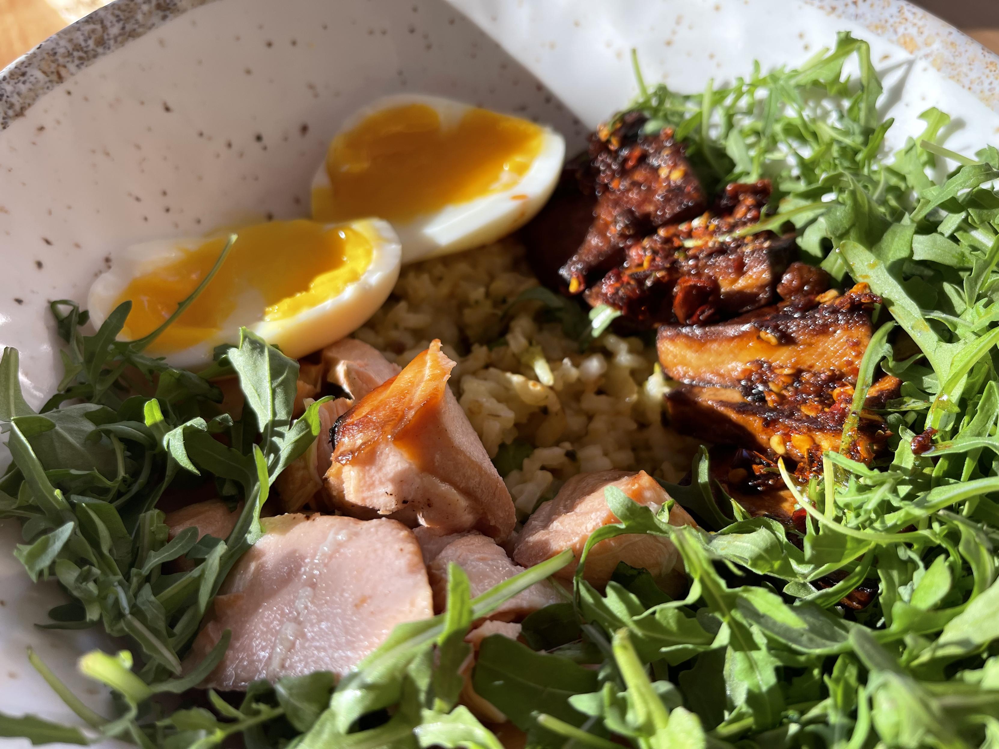 W misce znajduje się kremowe risotto z zielonym groszkiem. Na wierzchu ułożone są kawałki łososia, podsmażone tofu w przyprawach, rukola oraz jajko na półmiękko z płynnym żółtkiem.