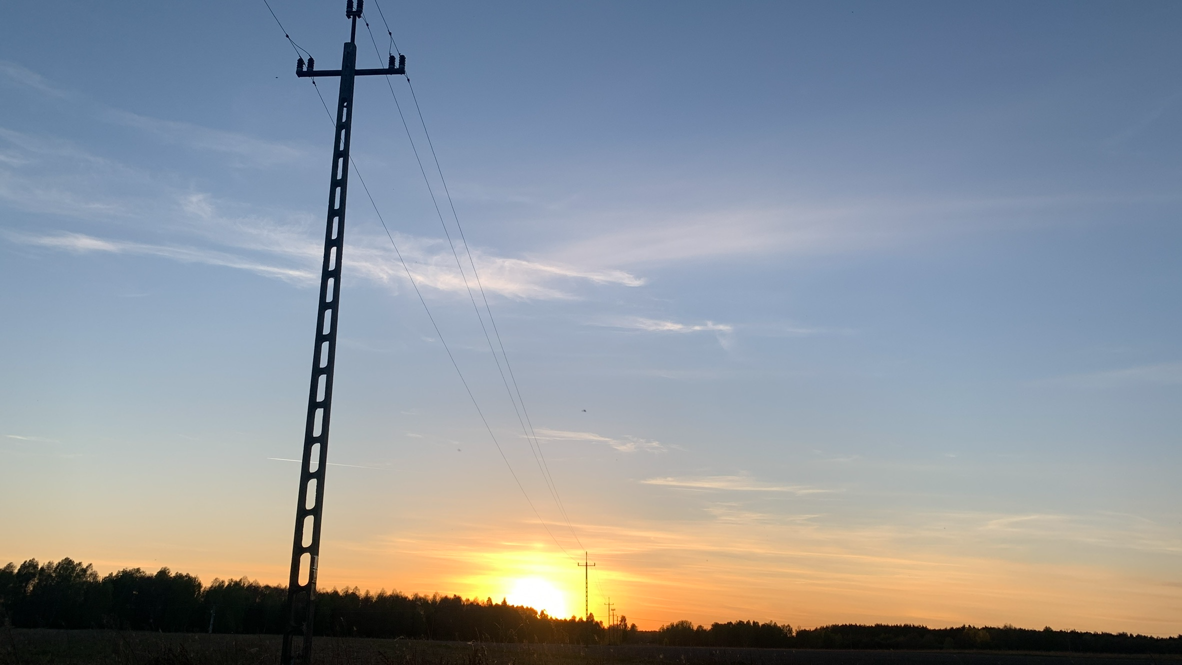 Słup energetyczny, a dalej kolejny i kolejne, coraz mniejsze. W tle zachodzące słońce nad lasem i polami
