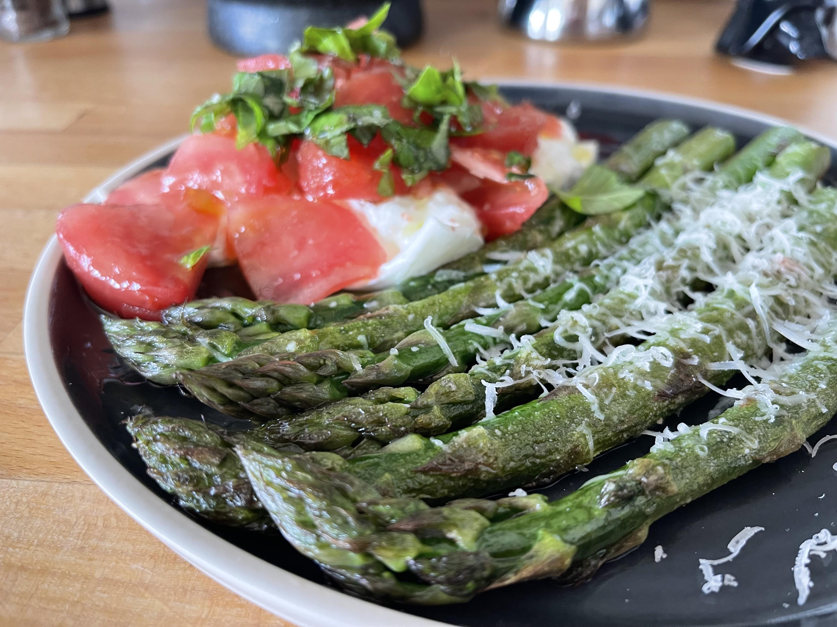 Na czarnym talerzu leżą grillowane zielone szparagi posypane startym parmezanem, obok znajduje się burrata przykryta kawałkami czerwonego pomidora malinowego i świeżą bazylią