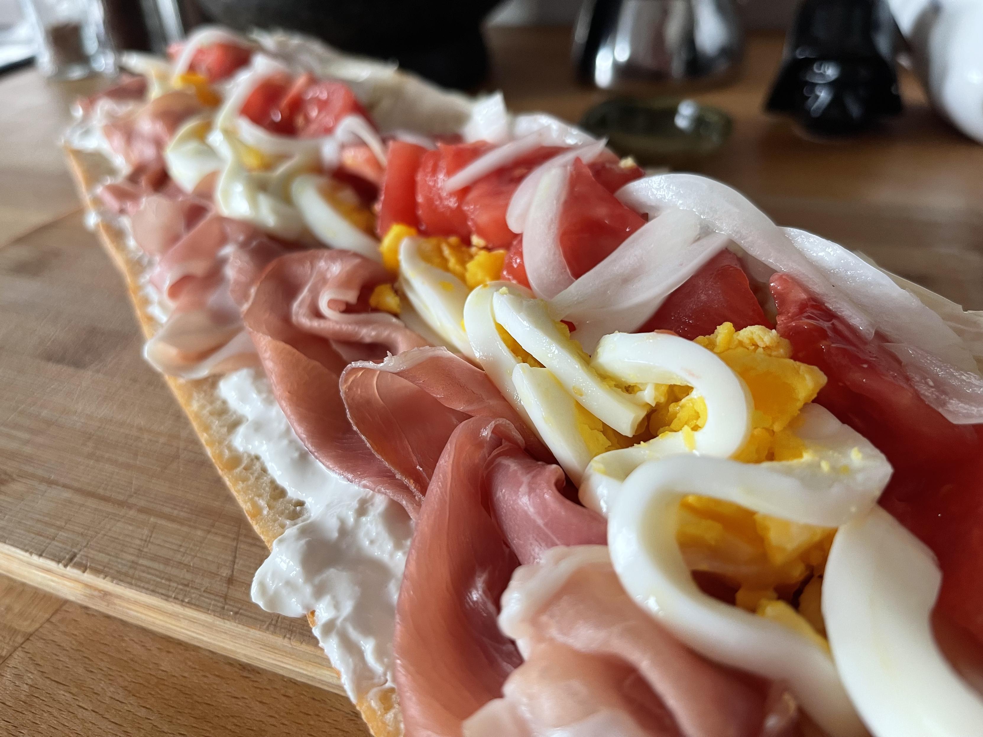 Na drewnianej desce leży chrupiąca bagietka posmarowana burratą i obłożona plastrami szynki serrano, kawałkami jajka na twardo, świeżym pomidorem oraz cienko krojoną cebulką.