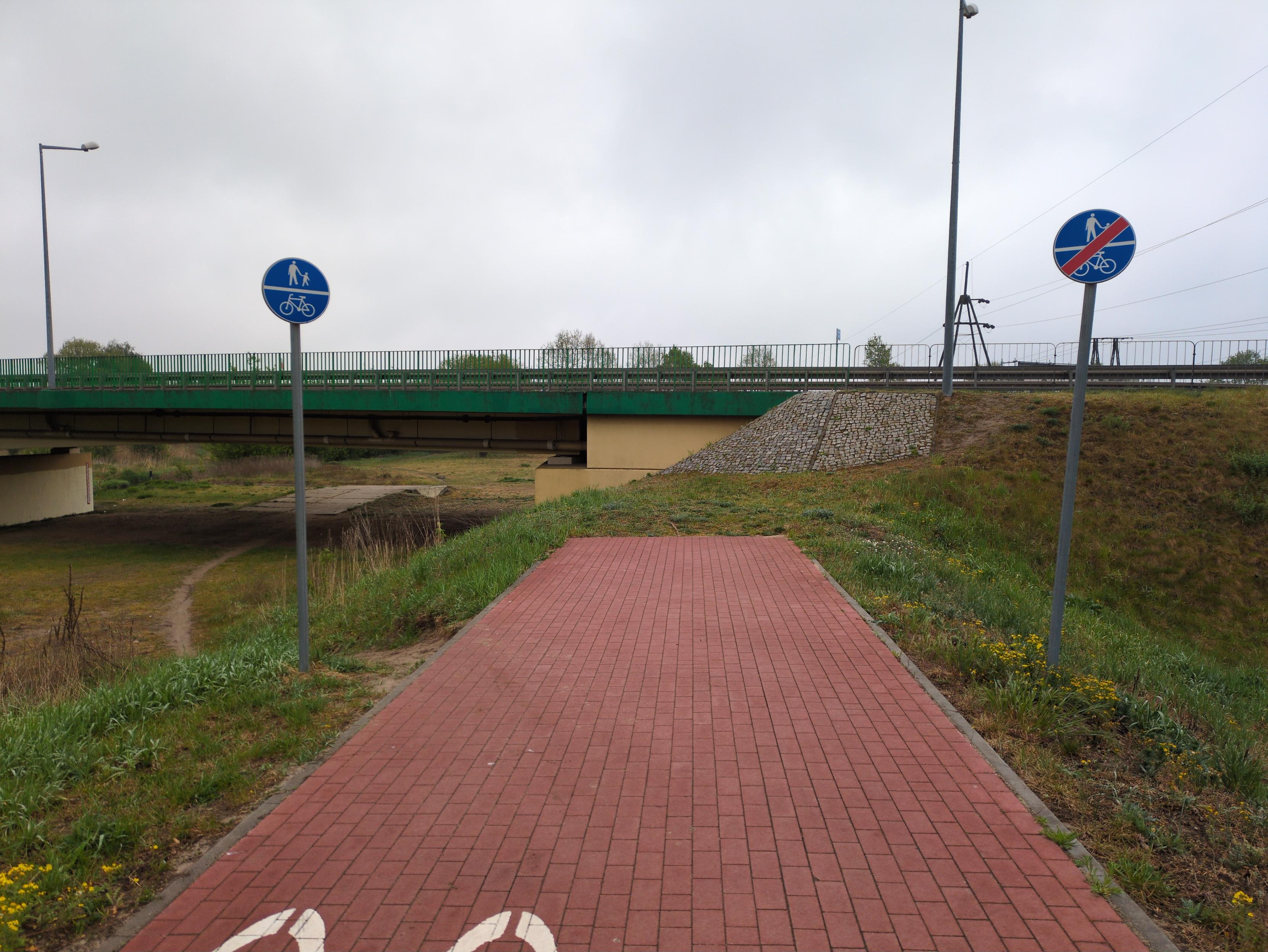 Zdjęcie przedstawiające drogę dla rowerów i pieszych z czerwonej kostki, na wale ziemnym. Droga nagle ucina się kawałek dalej, przed idącym w poprzek mostem. Most przebiega nieco wyżej, otoczony jest barierkami i nie ma opcji wejścia na niego. Drogę otacza trawa. Niedaleko przed końcem drogę otaczają po obu stronach znaki: z prawej obwieszczający koniec drogi dla pieszych i rowerów, z lewej na tej samej wysokości początek. Tuż za znakiem zaczyna się wydeptana w trawie ścieżka, który schodzi po zboczu i łukiem zakręca pod most. Kawałek dalej wchodzi w betonowe płyty.