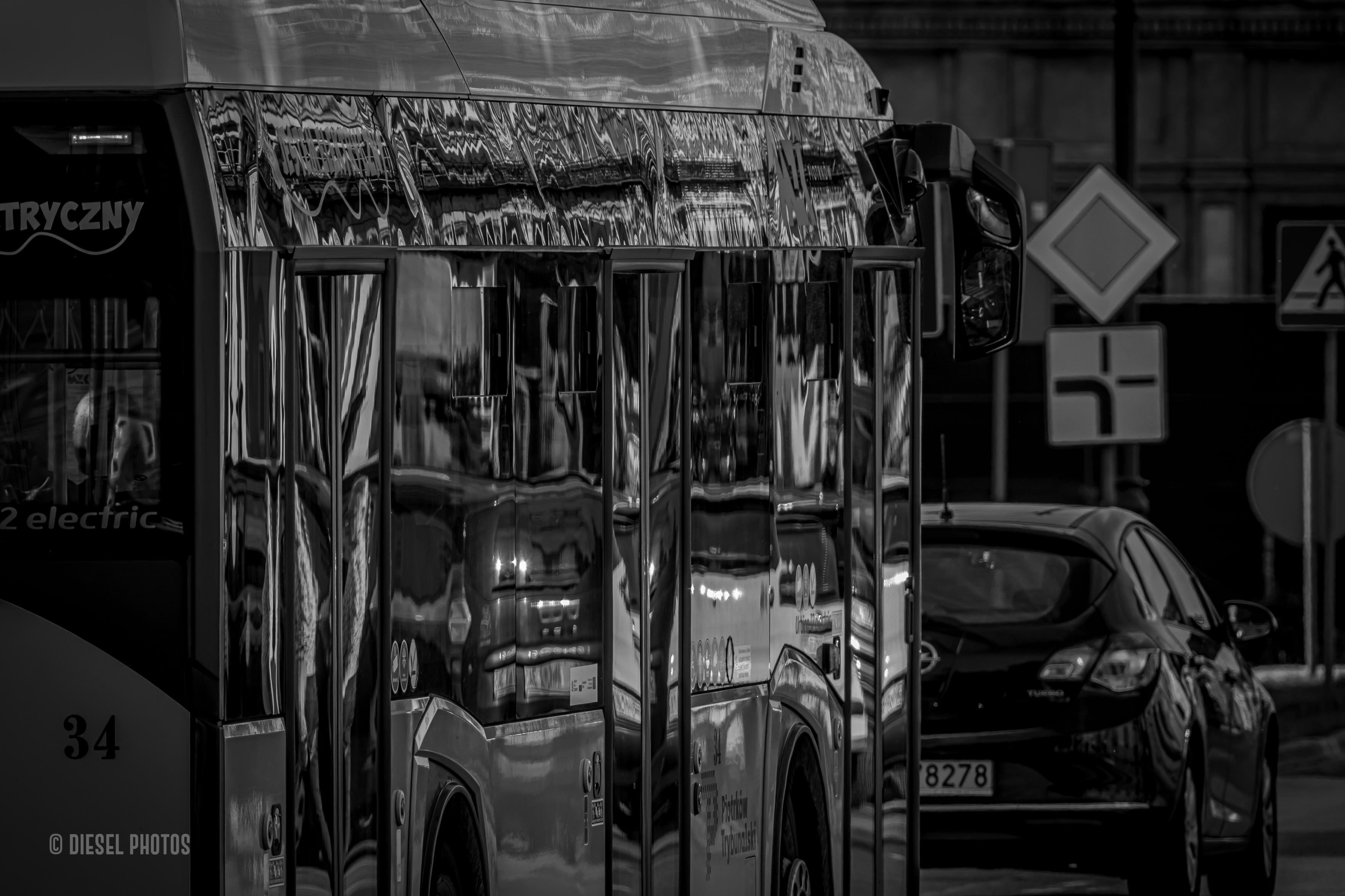 Czarno-białe zdjęcie przedstawia fragment nowoczeszego autobusu miejskiego sfotografowanego z boku, z niewielkiego kąta. Kadr skupia się niemal wyłącznie na jego błyszczących, mocno przyciemnionych szybach, które działają jak ogromne lustro.

W odbiciach szyb widać zdeformowany obraz otaczającego miasta – elewacje kamienic, samochody, światła i uliczną infrastrukturę. Pionowe słupki między oknami dzielą odbicia na wąskie pasy, przez co budynki i pojazdy wydają się pofalowane, jakby oglądane przez poruszoną taflę wody. Powstaje abstrakcyjna kompozycja złożona z pionowych linii, refleksów i zniekształconych kształtów.

Po prawej stronie kadru znajduje się ciemny samochód osobowy stojący lub powoli jadący przed autobusem. W tle widoczne są znaki drogowe o charakterystycznych geometrycznych kształtach, lekko rozmyte i odsunięte na drugi plan.

Całość utrzymana jest w mocnych kontrastach czerni, bieli i odcieni szarości. Brak koloru sprawia, że uwagę przyciąga przede wszystkim gra światła, odbić i rytmicznych pionowych podziałów szyb. Zdjęcie ma bardziej abstrakcyjny niż dokumentalny charakter – zamiast pokazywać sam autobus, koncentruje się na zniekształconym obrazie miasta odbijającym się w jego lustrzanej powierzchni.