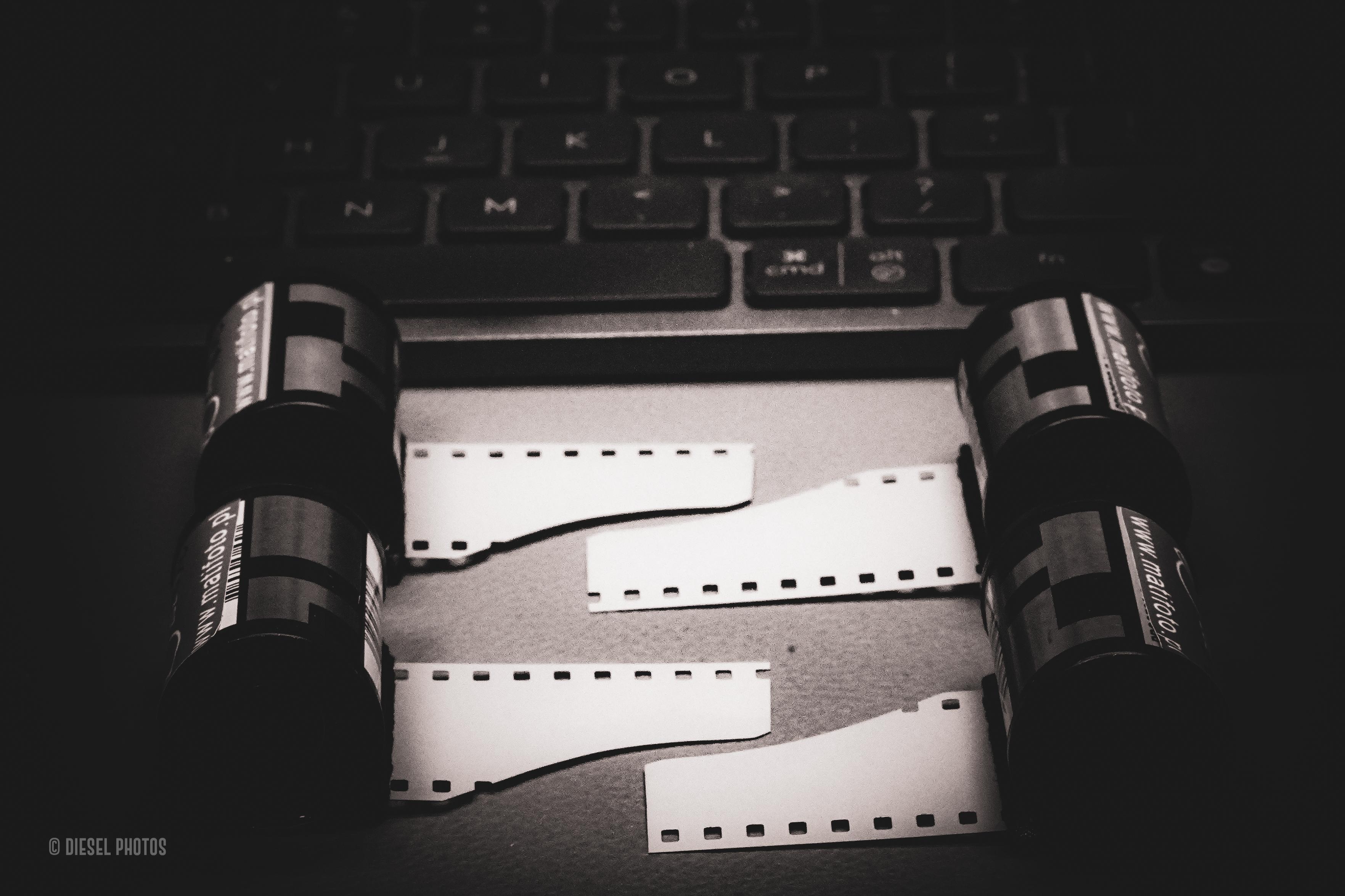 Czarno-białe zdjęcie przedstawia cztery pojemniki z filmem fotograficznym 35 mm ustawione po lewej i prawej stronie kadru. Pomiędzy nimi leżą cztery krótkie paski jasnego negatywu z wyraźnie widoczną perforacją. W tle znajduje się rozmyta czarna klawiatura komputerowa, tworząca kontrast między tradycyjną fotografią analogową a współczesnym, cyfrowym światem. Najjaśniejszym elementem zdjęcia są paski filmu, wyraźnie odcinające się od ciemnego tła. Całość utrzymana jest w minimalistycznej, spokojnej stylistyce.