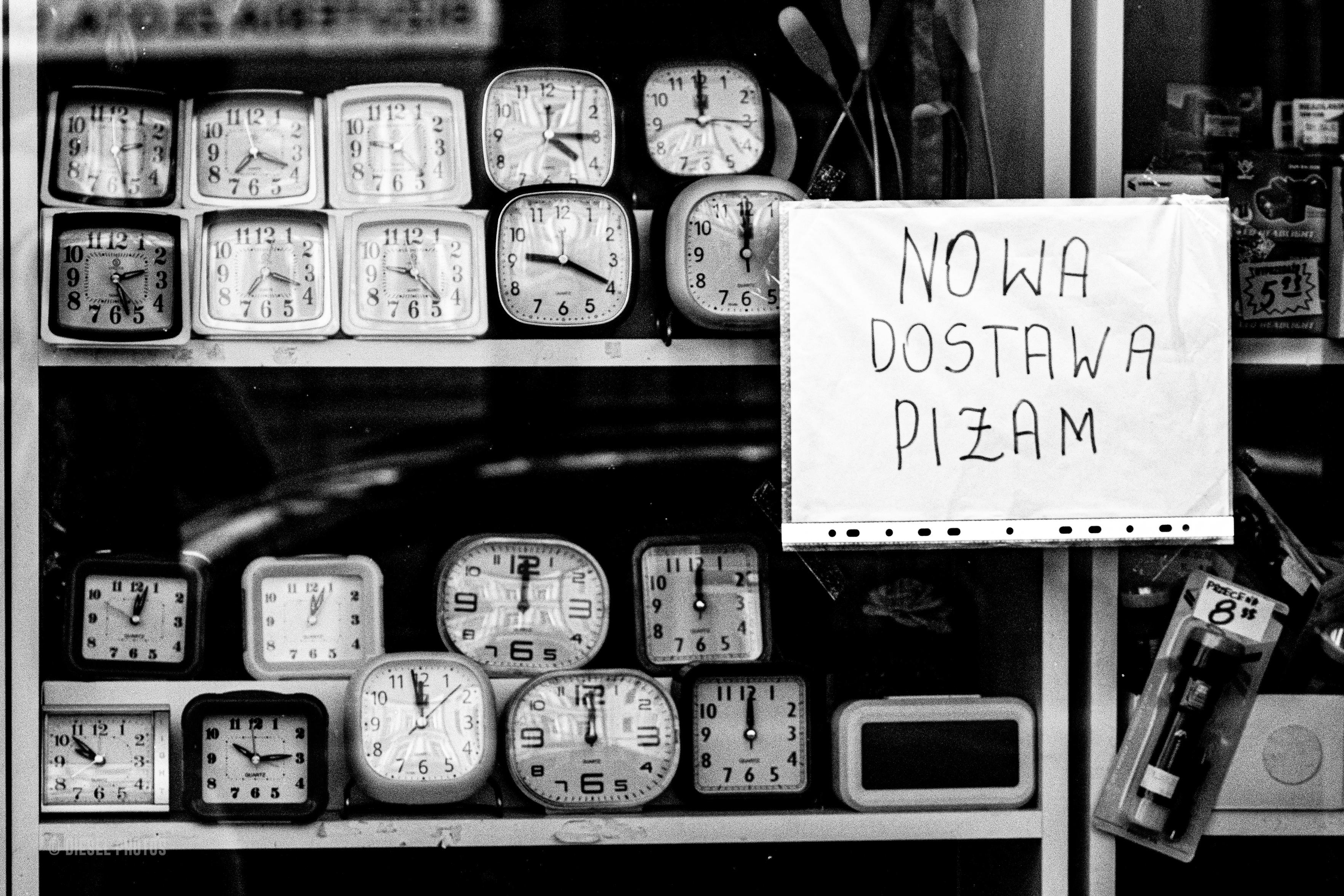 Czarno-białe zdjęcie przedstawia witrynę niewielkiego sklepu z kilkunastoma analogowymi budzikami ustawionymi na półkach. Większość z nich wskazuje godzinę dwunastą lub kilka minut po niej, choć część pokazuje inne godziny. Po prawej stronie wisi odręcznie napisany karton z napisem „NOWA DOSTAWA PIŻAM”. Obok widoczne są różne drobne towary, między innymi latarka w plastikowym opakowaniu. Zdjęcie zestawia świat odmierzania czasu z codziennością małego sklepu, tworząc prostą, nieco humorystyczną scenę uliczną.