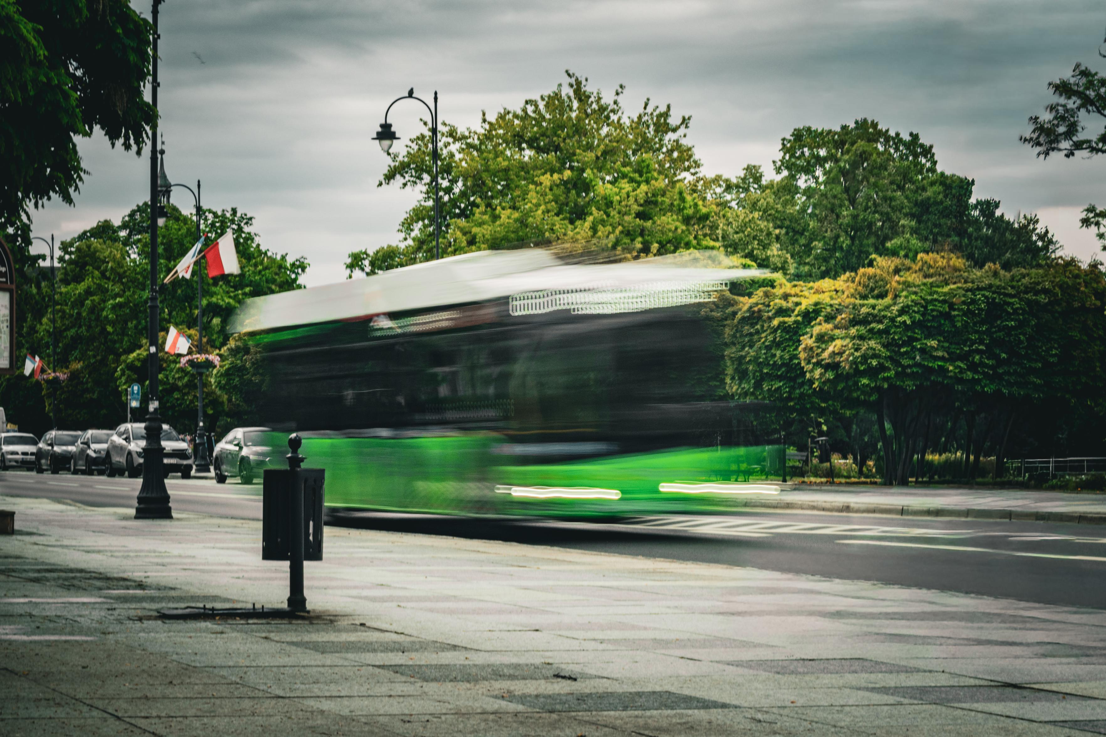 Kolorowe zdjęcie przedstawia zielony autobus miejski sfotografowany z długim czasem naświetlania, dzięki czemu jego sylwetka jest mocno rozmyta i sprawia wrażenie pędzącej przez kadr. Ostre pozostają jedynie otoczenie – mokra jezdnia, chodnik, zaparkowane samochody oraz drzewa w tle. Po lewej stronie ulicy wiszą biało-czerwone polskie flagi na latarniach, a nad całą sceną rozciąga się ciężkie, pochmurne niebo. Rozmyty autobus kontrastuje ze spokojnym, nieruchomym otoczeniem, podkreślając wrażenie ruchu i dynamiki.