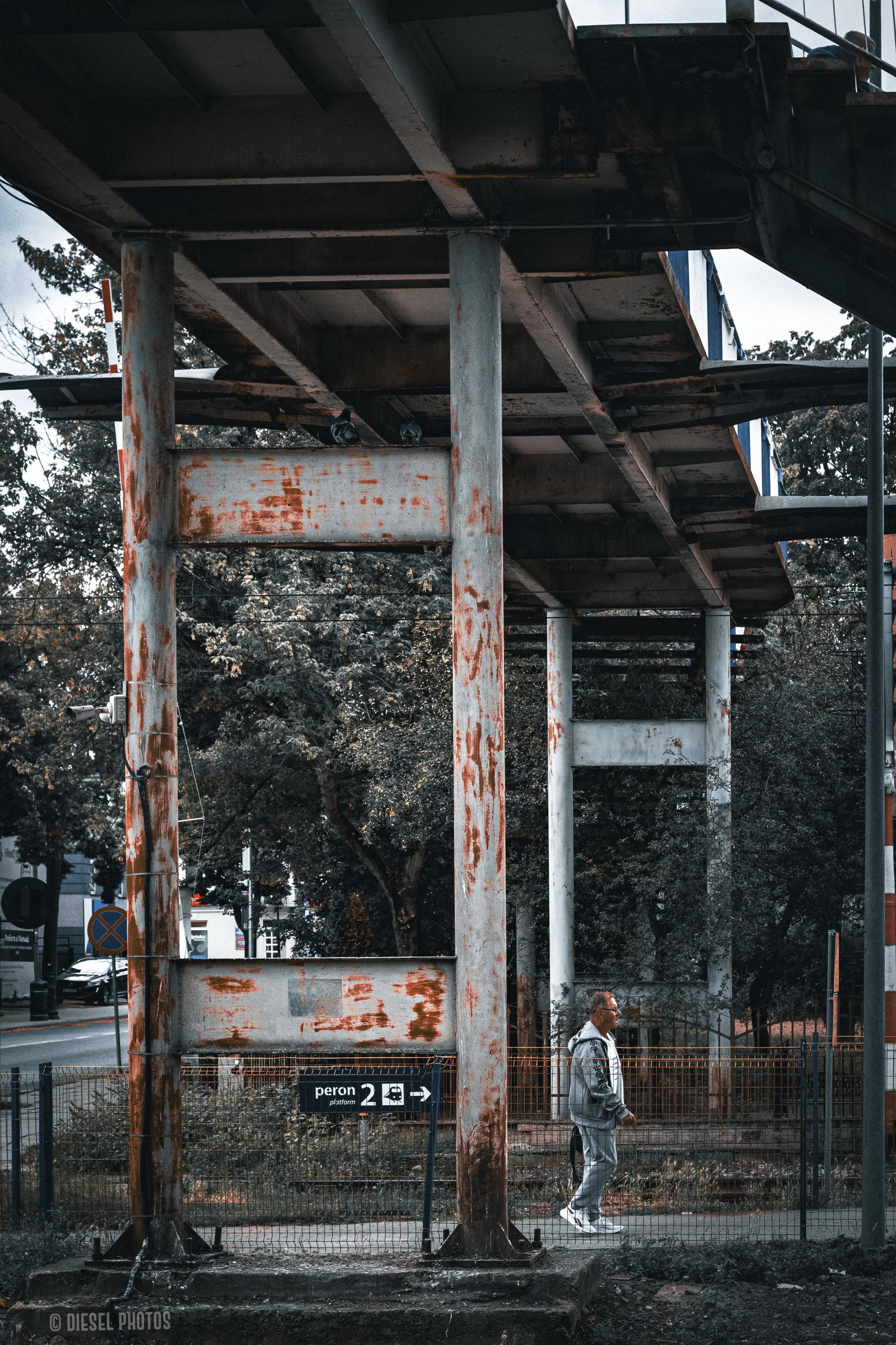 Metalowa konstrukcja zadaszenia peronu sfotografowana od boku. Grube, miejscami zardzewiałe słupy i belki tworzą rytmiczny układ prowadzący w głąb kadru. Między słupami, przy ogrodzeniu, idzie starszy mężczyzna w jasnym dresie i białych butach. Na pierwszym planie widoczna jest tabliczka z napisem „Peron 2”. Tło wypełniają drzewa oraz fragment ulicy. Zdjęcie utrzymane jest w chłodnych, przygaszonych barwach z wyraźnie podkreśloną rdzą na stalowej konstrukcji.