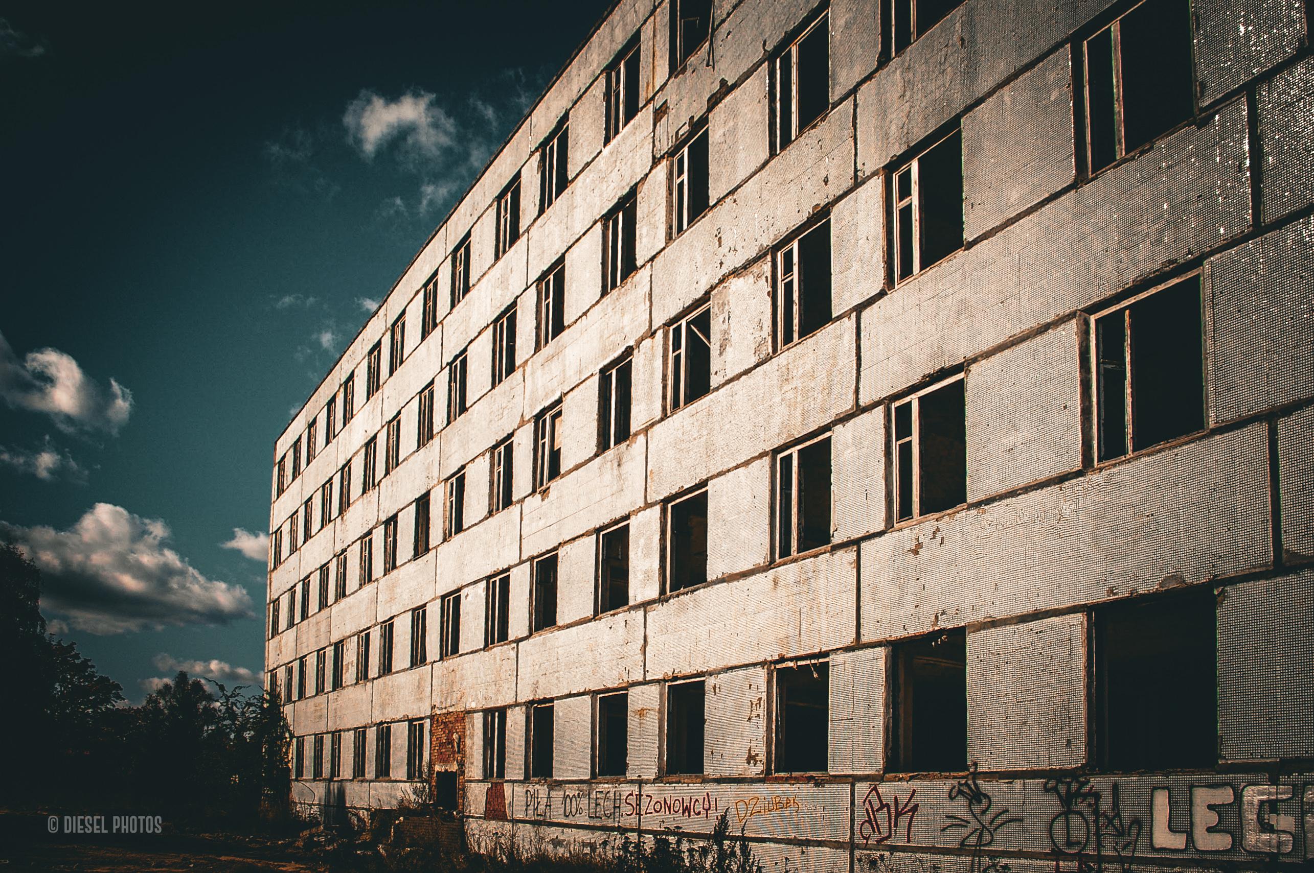 Na zdjęciu widać długi, pięciopiętrowy blok z wielkiej płyty sfotografowany z niskiej perspektywy. Budynek ciągnie się ukośnie przez niemal cały kadr, dzięki czemu wydaje się bardzo długi i masywny. Większość okien jest pozbawiona szyb, a ciemne otwory kontrastują z jasną, mocno zużytą elewacją. Na parterze widoczne są graffiti i ślady zniszczeń, a wokół wyrasta dzika roślinność. Po lewej stronie znajduje się ciemne niebo z kilkoma jasnymi chmurami. Całość utrzymana jest w ciepłych, lekko brązowych tonach, co podkreśla opuszczony, postapokaliptyczny charakter miejsca.