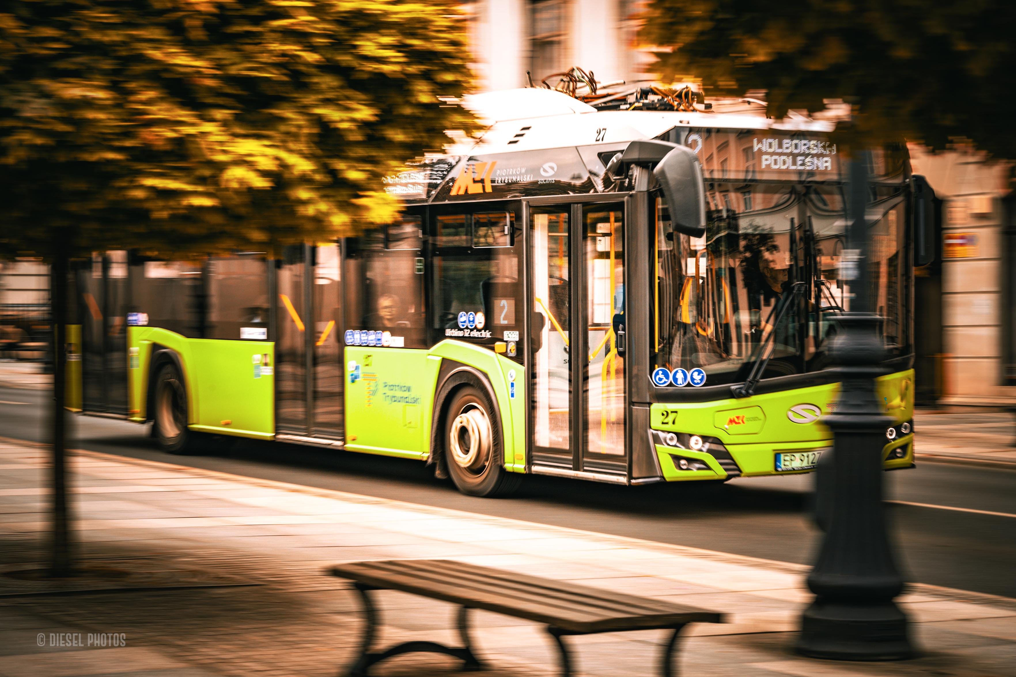 Nowoczesny zielony autobus komunikacji miejskiej jedzie ulicą w Piotrkowie Trybunalskim. Zastosowane długie naświetlanie i płynny ruch aparatu sprawiają, że pojazd pozostaje wyraźny, podczas gdy drzewa, chodnik i budynki rozmywają się w kierunku jazdy, podkreślając wrażenie prędkości. Na elektronicznym wyświetlaczu widoczny jest kierunek „Wolborska Podleśna”. Na pierwszym planie znajduje się częściowo rozmyta ławka oraz latarnia, które tworzą dodatkową głębię kadru. Całość utrzymana jest w ciepłej kolorystyce, kontrastującej z intensywną zielenią autobusu.