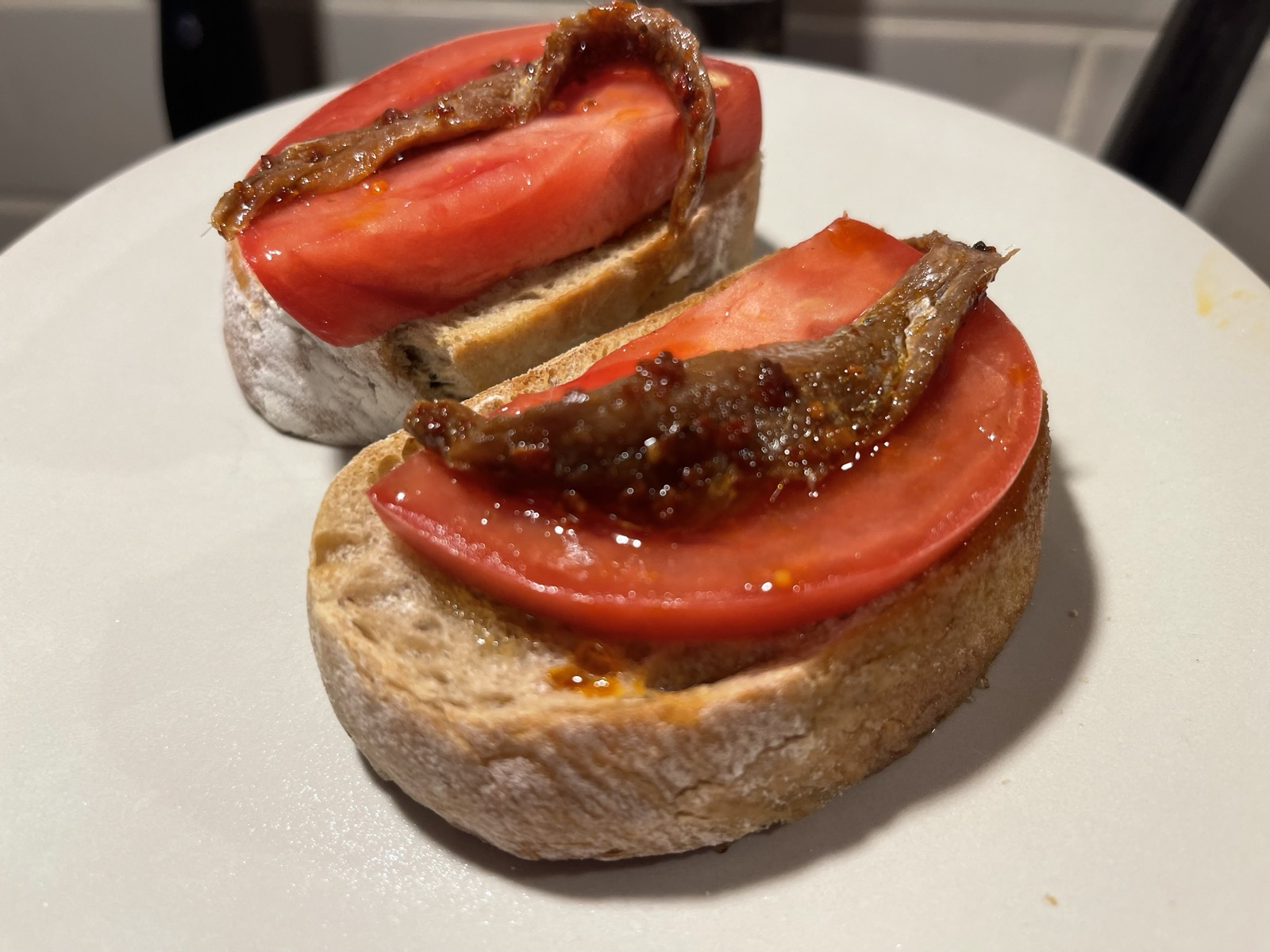 Dwie kromki jasnej bagietki ułożone na białym talerzu. Na każdej znajduje się gruby plaster dojrzałego, czerwonego pomidora oraz ciemny filet anchois lub sardeli, pokryty błyszczącą oliwą z drobnymi kawałkami przypraw. Zdjęcie wykonano z bliska, na tle rozmytej kuchni.