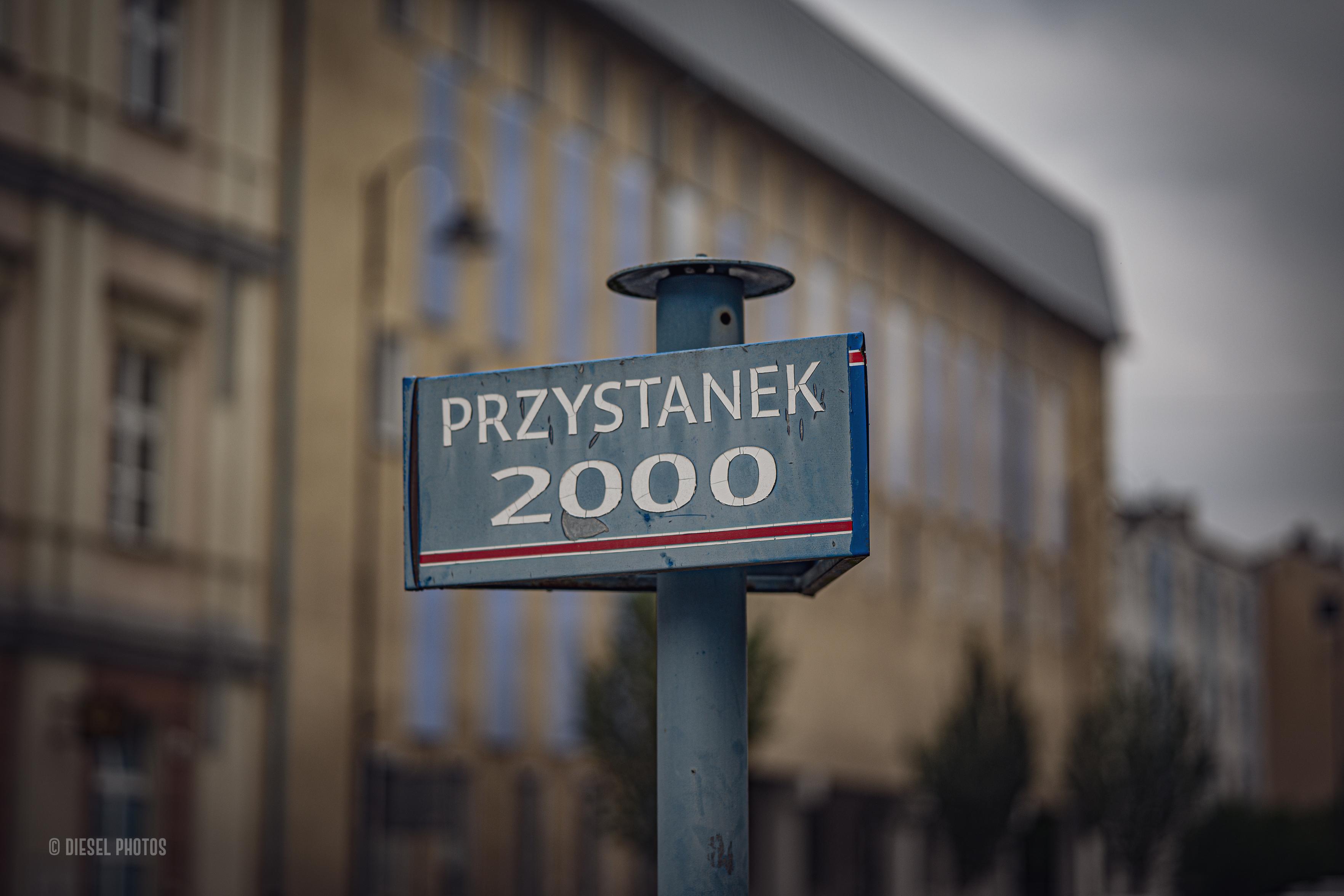 Na pierwszym planie znajduje się stary, niebieski znak przystankowy z napisem „PRZYSTANEK 2000”. Farba jest miejscami wytarta, a tabliczka nosi ślady wieloletniego użytkowania. Dzięki niewielkiej głębi ostrości znak pozostaje wyraźny, podczas gdy tło miękko się rozmywa. Za nim widać fasadę zabytkowej kamienicy z wysokimi oknami i jasnym tynkiem oraz pochmurne, szare niebo. Minimalistyczna kompozycja skupia uwagę na prostym miejskim detalu, który przywołuje skojarzenia z początkiem nowego tysiąclecia i dawnym wyglądem polskich ulic.