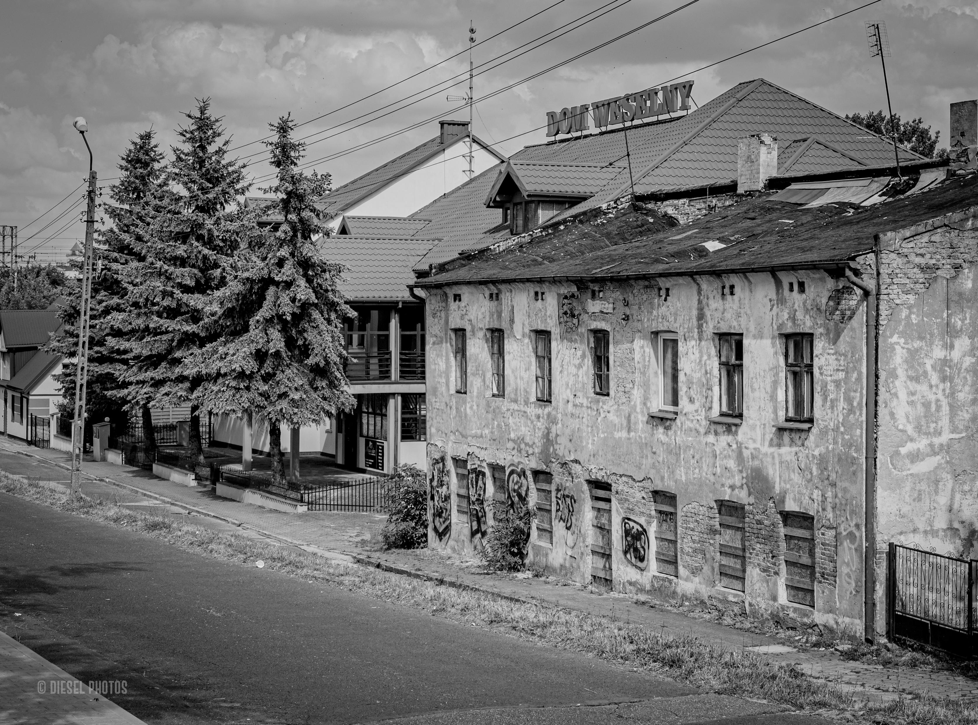 Czarno-biała fotografia przedstawia długi, mocno zniszczony, parterowy budynek stojący przy spokojnej ulicy. Elewacja jest popękana, miejscami odpada tynk, odsłaniając cegły. Dolna część ściany pokryta jest graffiti, a większość okien wygląda na opuszczone lub zabezpieczone.

Za starym budynkiem widać nowoczesne zabudowania z jasnymi elewacjami i zadbanymi dachami. Na jednym z nich znajduje się duży napis „Dom Weselny”, górujący nad okolicą.

Po lewej stronie rosną wysokie świerki, a wzdłuż ulicy biegnie chodnik oraz słupy z przewodami energetycznymi. Nad całą sceną wiszą ciężkie, warstwowe chmury, które nadają fotografii spokojny, lekko melancholijny charakter.