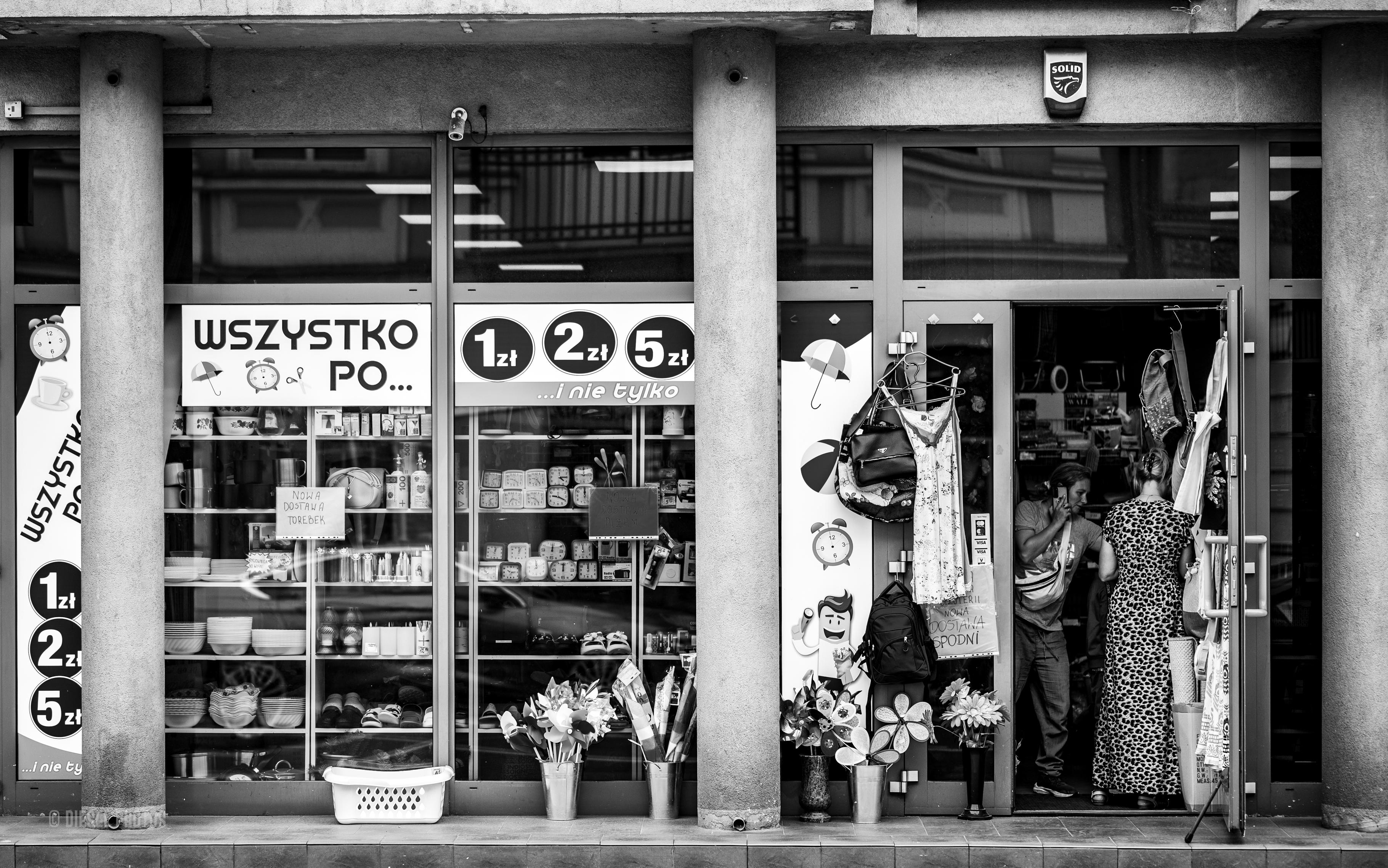 Na fotografii widać niewielki sklep typu „wszystko po…”, którego witryna wypełniona jest drobnymi przedmiotami codziennego użytku. Za szybą stoją naczynia, zegary, kosmetyki, dekoracje i inne niedrogie artykuły, a duże napisy „1 zł”, „2 zł” i „5 zł” przyciągają wzrok przechodniów. Przy wejściu wiszą torebki i ubrania, obok ustawiono sztuczne kwiaty w metalowych wiadrach. W drzwiach dwie kobiety prowadzą rozmowę, tworząc naturalną, codzienną scenę z życia miasta. Czarno-biała obróbka podkreśla rytm betonowych kolumn, bogactwo detali witryny oraz kontrast między uporządkowaną fasadą a zwyczajnością niewielkiego osiedlowego handlu.