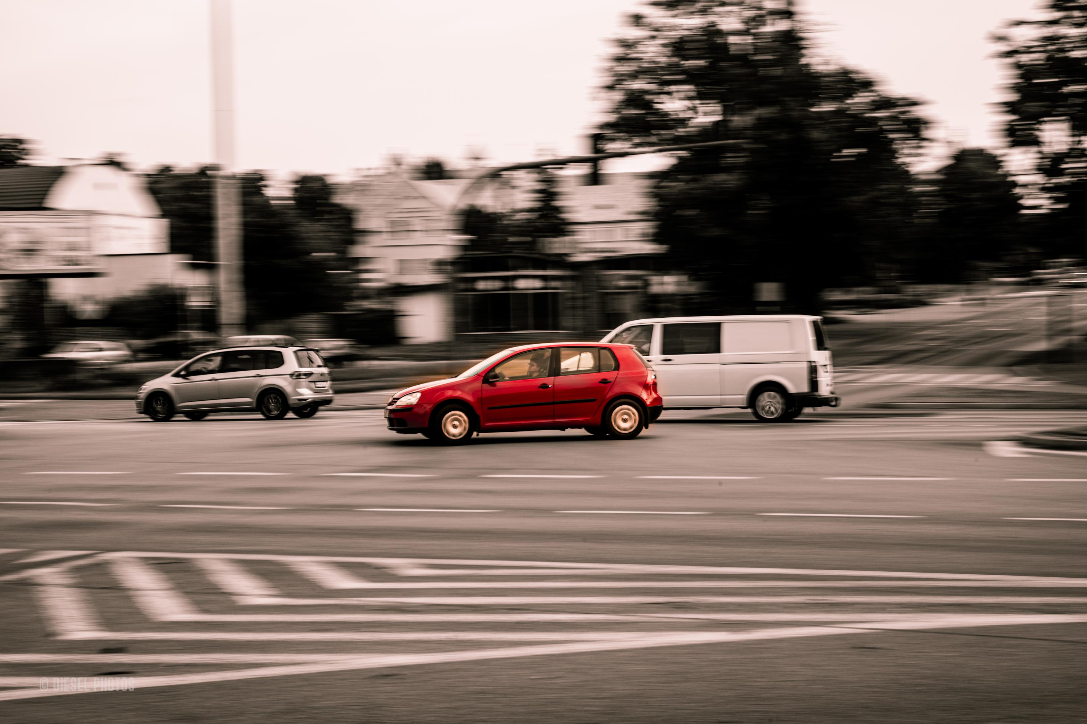 Na zdjęciu widać ruchliwe miejskie skrzyżowanie sfotografowane techniką panoramowania. W centrum kadru znajduje się czerwony, pięciodrzwiowy hatchback jadący z lewej strony na prawą. Samochód jest stosunkowo ostry, podczas gdy tło – budynki, drzewa i jezdnia – zostało poziomo rozmyte, co wyraźnie podkreśla wrażenie prędkości.

Pozostałe pojazdy, srebrny samochód po lewej i biały dostawczy van po prawej, są mniej wyeksponowane i wtapiają się w rozmyte otoczenie. Cała scena utrzymana jest w odcieniach czerni, bieli i sepii, natomiast czerwony samochód zachował swój intensywny kolor, dzięki czemu natychmiast przyciąga wzrok.

Na pierwszym planie widoczne są pasy i oznakowanie jezdni, również lekko rozciągnięte przez ruch aparatu. W tle majaczą drzewa, domy i sygnalizacja świetlna, ale pozostają jedynie sugestią miejskiej przestrzeni. Zdjęcie sprawia wrażenie uchwycenia krótkiej chwili w nieustannym ruchu ulicznego życia, a kontrast między ostrym samochodem i rozmytym otoczeniem buduje silne poczucie dynamiki.