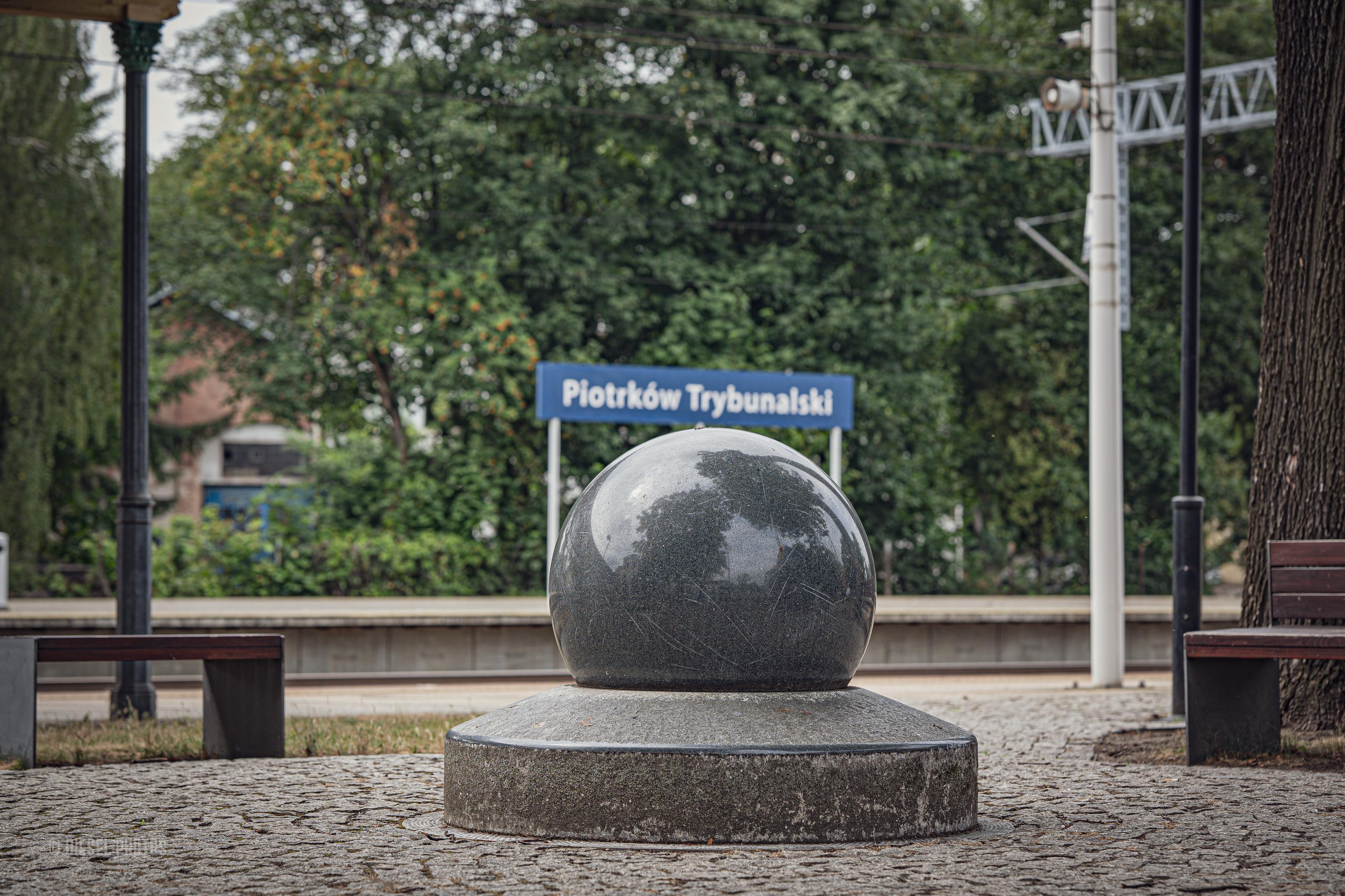 Na zdjęciu w centrum kadru znajduje się duża, wypolerowana kamienna kula osadzona na okrągłym cokole. Ustawiono ją na placu wyłożonym kostką brukową, a fotograf wykonał zdjęcie dokładnie na wprost, dzięki czemu kompozycja jest niemal idealnie symetryczna.

Za kulą, lekko rozmyty, widoczny jest niebieski peronowy znak z napisem „Piotrków Trybunalski”, wskazujący lokalizację dworca kolejowego. Po obu stronach kadru stoją ławki i wysokie drzewa, a w tle widać perony, słupy trakcji kolejowej oraz gęstą zieleń. Na błyszczącej powierzchni kamiennej kuli odbijają się korony drzew i fragment nieba.

Całość utrzymana jest w spokojnej, stonowanej kolorystyce. Zdjęcie sprawia wrażenie uporządkowanego i statycznego, bardziej przypominając studium geometrii i symetrii niż klasyczny reportaż z dworca.