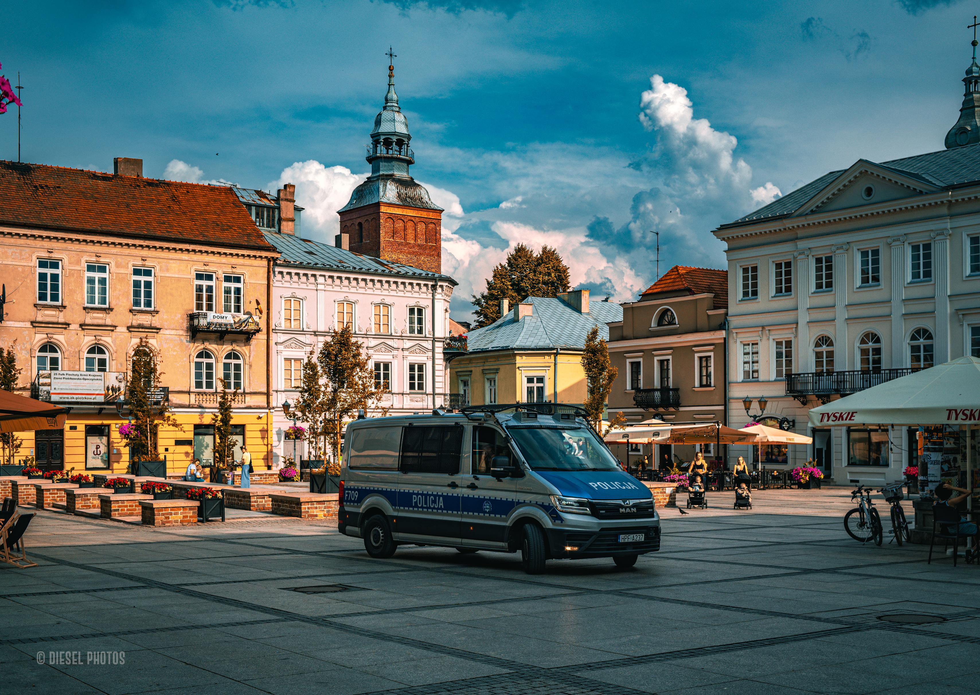 Na fotografii przedstawiono rynek w Piotrkowie Trybunalskim w ciepłym, późnopopołudniowym świetle. W centrum kadru przez rozległy plac przejeżdża granatowo-srebrny radiowóz policyjny typu van, który kontrastuje z jasną nawierzchnią rynku i pastelowymi fasadami otaczających go kamienic. W tle wyrasta ceglana wieża kościoła z charakterystycznym hełmem, dominująca nad miejską zabudową. Po prawej stronie widać ogródki gastronomiczne, rowery oraz spacerujących mieszkańców i rodziny z wózkami, nadające scenie codzienny, spokojny charakter. Nad miastem rozciąga się efektowne niebo z wysokimi, białymi chmurami i ciemniejszymi pasmami błękitu, które wzmacniają kontrast z ciepłymi kolorami budynków. Zdjęcie łączy klasyczną architekturę historycznego centrum z elementami współczesnego życia, pokazując zwykły moment z miejskiej codzienności.