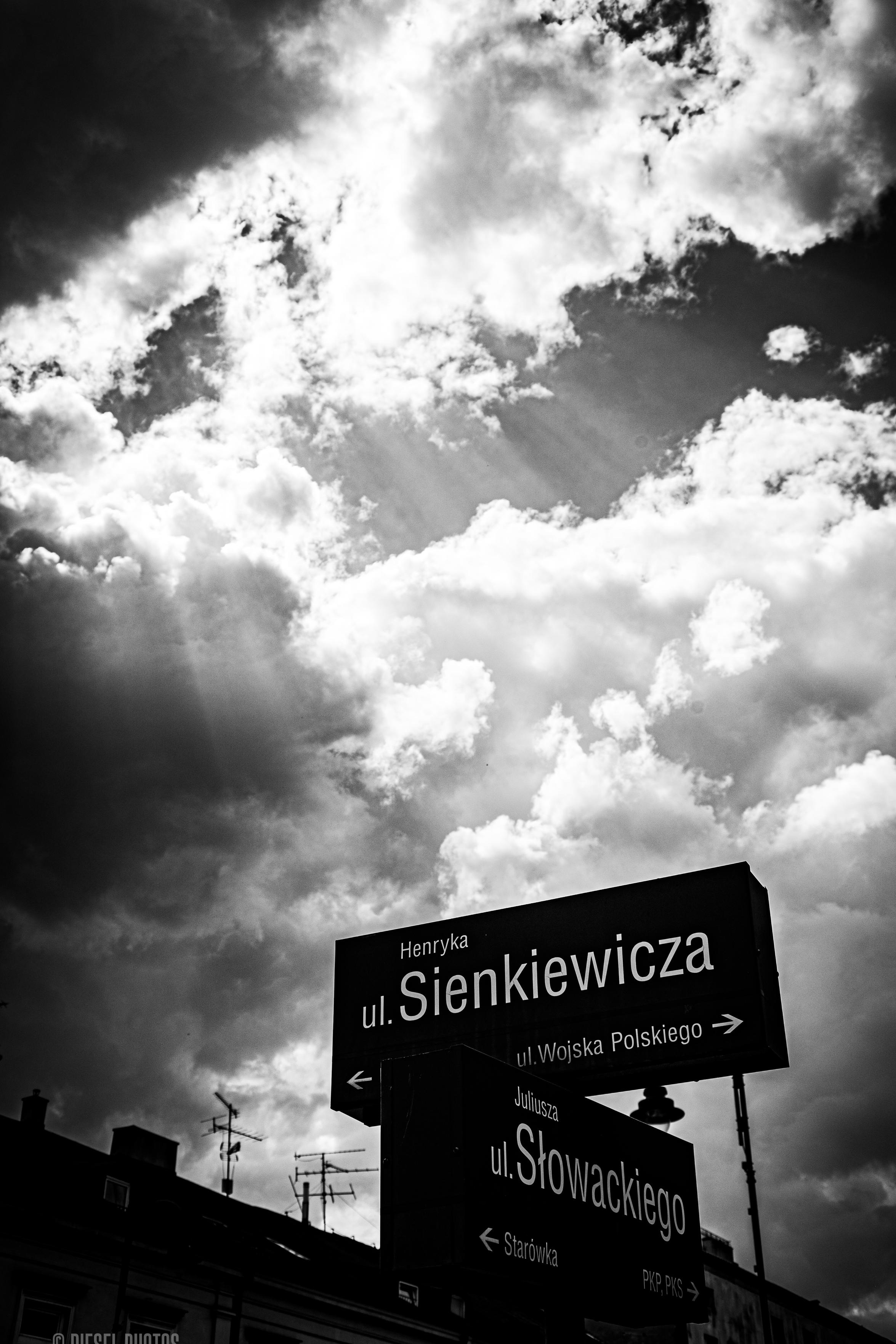 Czarno-biała fotografia wykonana z poziomu ulicy, skierowana wysoko ku niebu. Większość kadru zajmują gęste, burzowe chmury, pomiędzy którymi przebijają się jasne promienie słońca. W dolnej części zdjęcia znajdują się dwa miejskie drogowskazy ustawione jeden nad drugim. Górny wskazuje ulicę Henryka Sienkiewicza oraz kierunek do ulicy Wojska Polskiego, dolny – ulicę Juliusza Słowackiego i Stare Miasto. Ciemne tablice z białymi napisami wyraźnie odcinają się od jasnego nieba. Na samym dole widoczne są dachy kamienic z antenami telewizyjnymi. Całość ma mocny kontrast i dramatyczny, niemal filmowy nastrój.