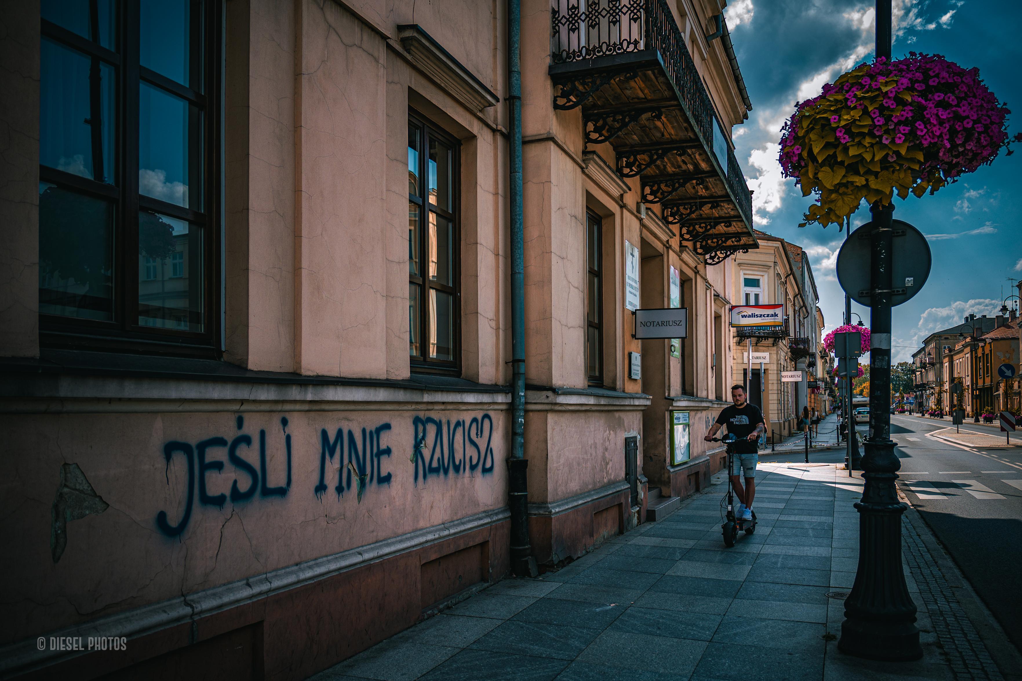 Kolorowa fotografia przedstawia fragment miejskiej ulicy z zabytkowymi kamienicami. Po lewej stronie kadru znajduje się jasna elewacja budynku, na której niebieską farbą sprayem namalowano duży napis: „JEŚLI MNIE RZUCISZ”. Napis urywa się i nie wiadomo, co miało znaleźć się dalej.

Środkiem chodnika jedzie na elektrycznej hulajnodze młody mężczyzna ubrany w ciemny T-shirt i jasne spodenki. Patrzy przed siebie i spokojnie mija budynki. Nad chodnikiem wiszą ozdobne balkony z kutymi balustradami, a przy latarniach zawieszono duże kosze pełne fioletowych kwiatów.

Po prawej stronie rozciąga się spokojna ulica z niewielkim ruchem samochodowym. W oddali widać kolejne kamienice i ciemne chmury na niebie, pomiędzy którymi przebija błękit. Zdjęcie zestawia elegancką architekturę z nieformalnym graffiti, tworząc kadr z lekkim poczuciem niedopowiedzenia i miejskiej ironii.