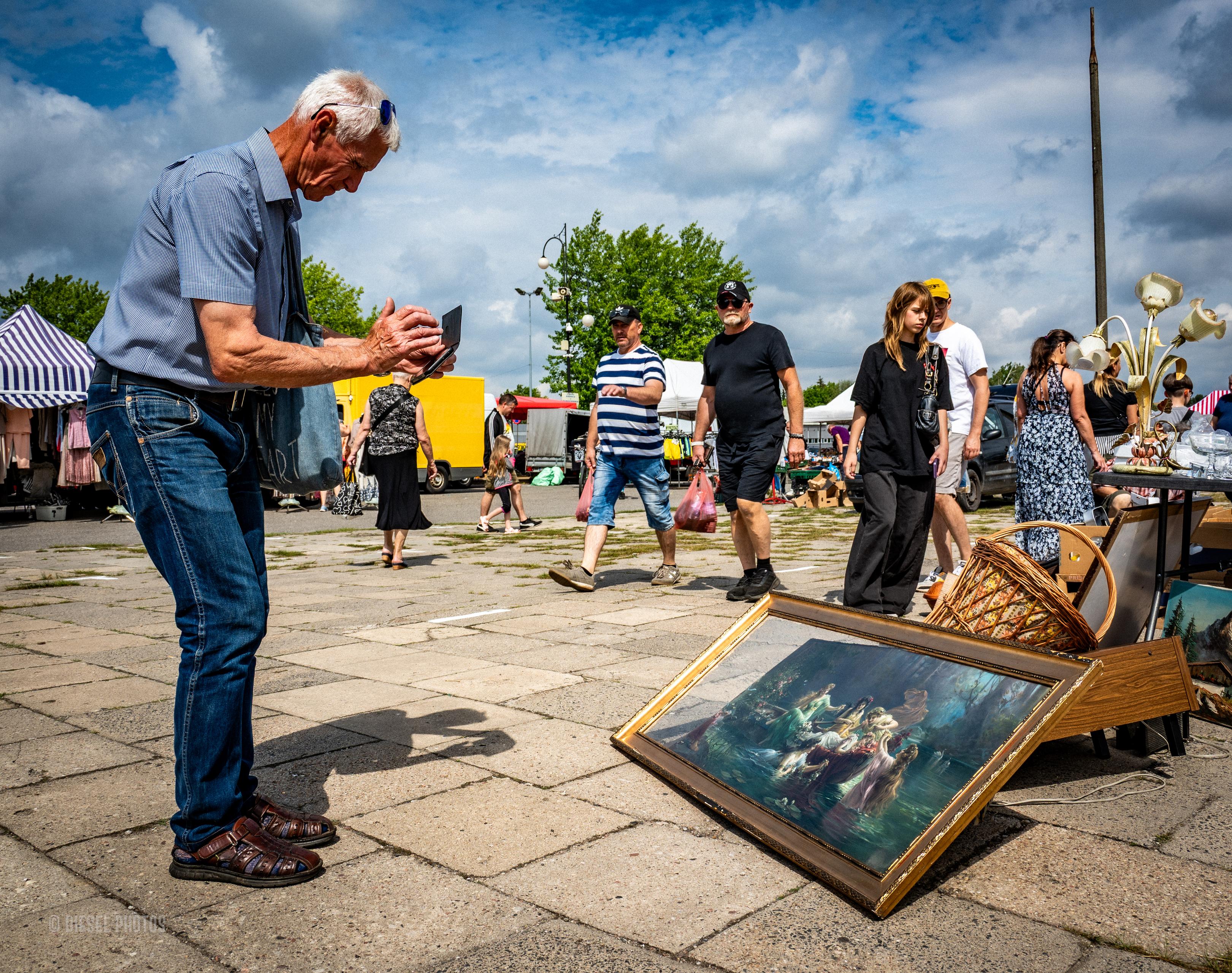 Kolorowa fotografia przedstawia scenę na targu staroci odbywającym się na otwartym placu. Po lewej stronie starszy mężczyzna z siwymi włosami pochyla się nad dużym obrazem opartym o ziemię. Trzyma w dłoniach smartfon lub niewielki tablet i fotografuje dzieło z bliskiej odległości. Ubrany jest w niebieską koszulę z krótkim rękawem, dżinsy i skórzane sandały.

Na pierwszym planie leży duży obraz w złotej ramie przedstawiający romantyczną scenę z kilkoma postaciami nad wodą. Obok stoją inne przedmioty wystawione na sprzedaż, między innymi wiklinowy kosz i stara lampa z kilkoma kloszami.

W tle widać spacerujących ludzi, stoiska targowe oraz błękitne niebo z dużymi, białymi chmurami. Zdjęcie sprawia wrażenie uchwycenia krótkiej, codziennej chwili, w której współczesna technologia spotyka się z  malarstwem.