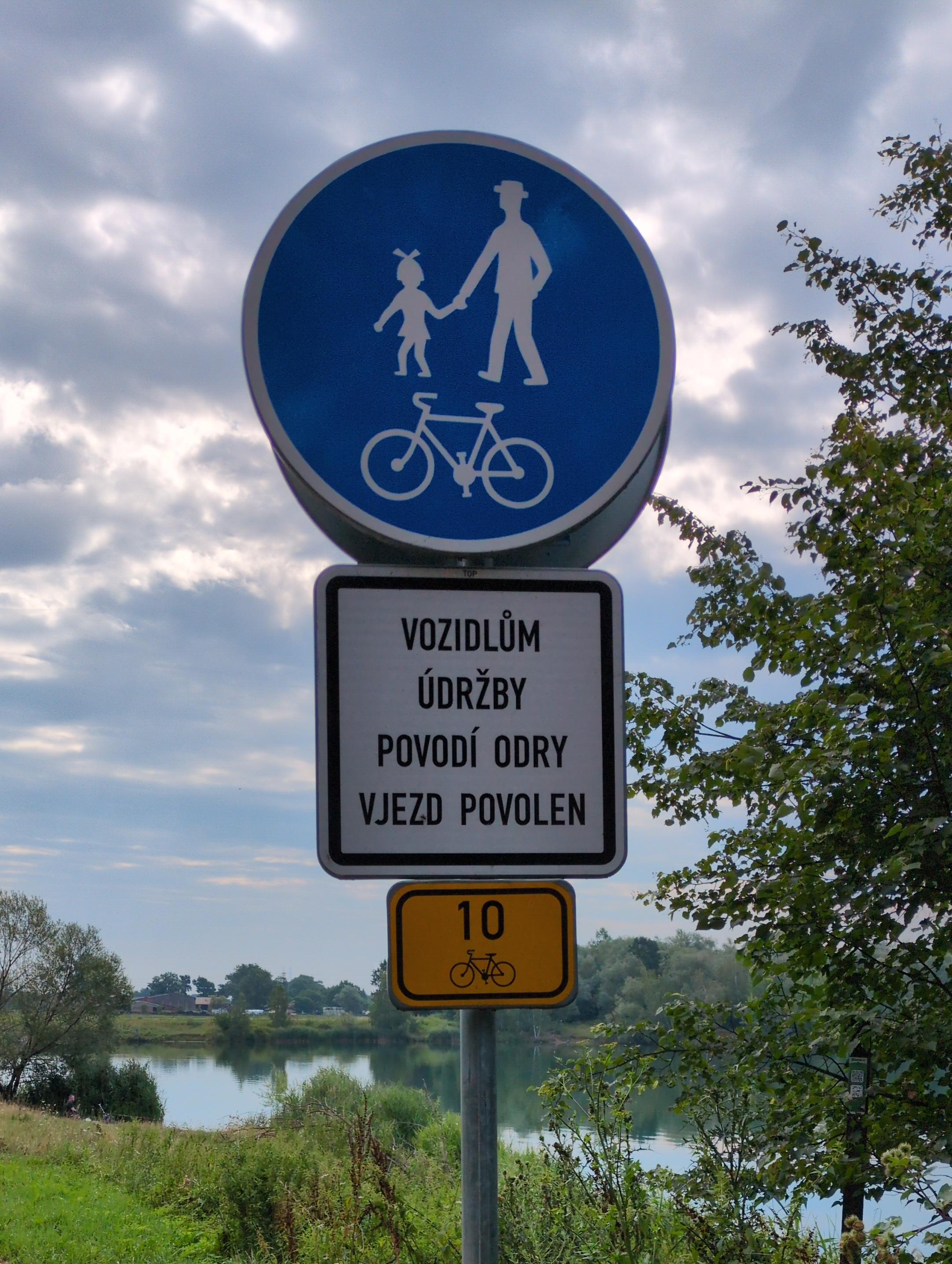 Trzyczęściowy znak drogowy. U góry w niebieskim kole z białą obwódką piktogram przedstawiający pana w kapeluszu, trzymającego za rękę dziewczynkę w krótkiej sukience, z kokardą na włosach, a poniżej rysunek roweru.

Pośrodku biały prostokąt z czarną obwódką, na którym napisane jest "vozidlům údržby povodí Odry vjezd povolen".

Na dole żółty prostokąt z czarną obwódką, a na nim liczba 10, pod nią mały rysunek roweru.