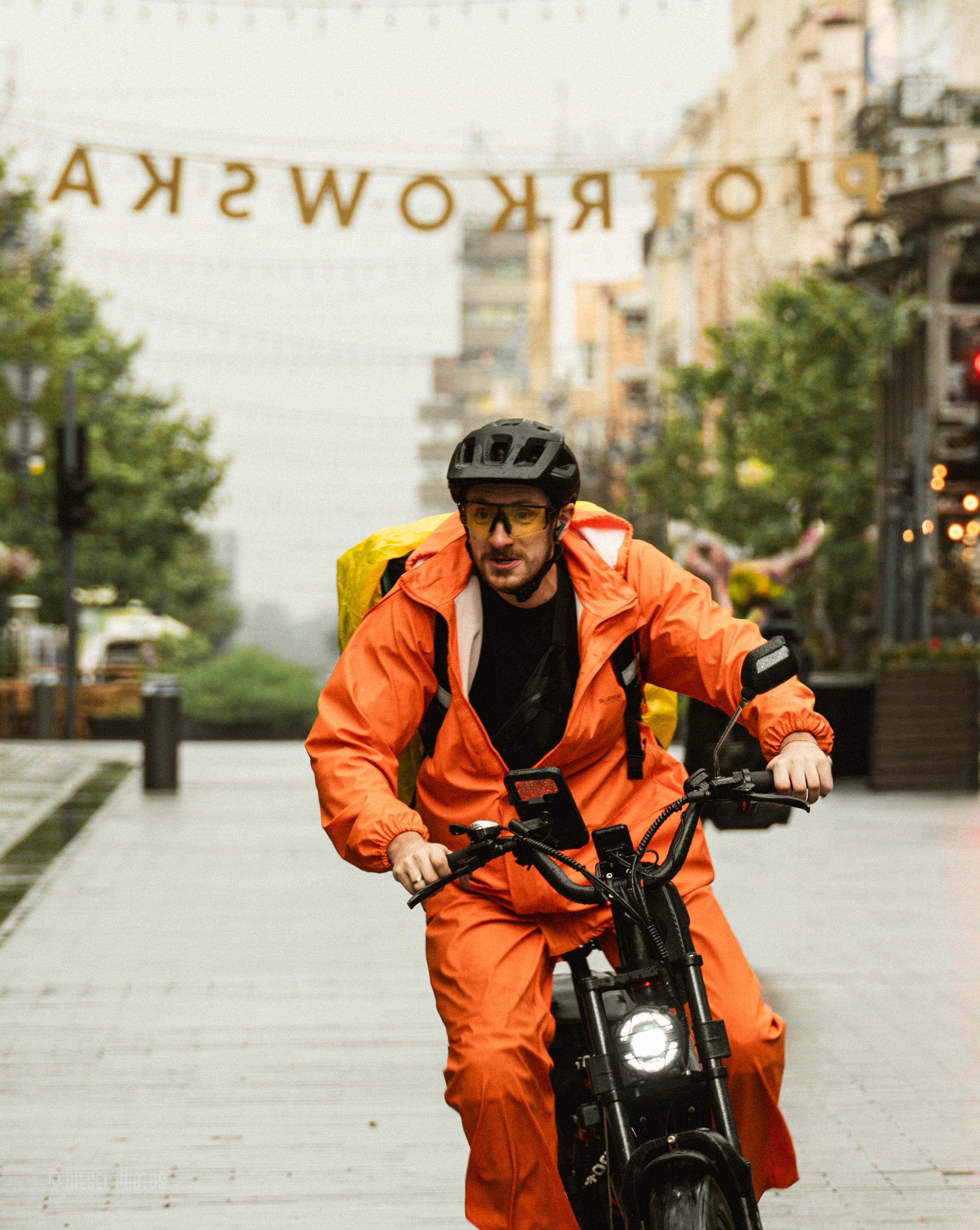 Na pierwszym planie ulicy jedzie rowerzysta na elektrycznym rowerze, ubrany w jaskrawopomarańczowy przeciwdeszczowy strój z dużym żółtym plecakiem kurierskim na plecach. Ma na głowie czarny kask i żółte okulary ochronne, a jego sylwetka jest ostra i wyraźna, co podkreśla wrażenie ruchu. Tło pozostaje miękko rozmyte – widać deptak obsadzony drzewami, latarnie, budynki i zamgloną perspektywę miasta po deszczu. Nad ulicą rozwieszony jest napis „PIOTRKOWSKA”, widoczny od tyłu, ponieważ fotografia została wykonana z przeciwnej strony. Mokra nawierzchnia odbija światło, a ciepły pomarańcz stroju kuriera mocno kontrastuje z chłodnymi szarościami i zielenią otoczenia, stając się dominującym elementem całej kompozycji.