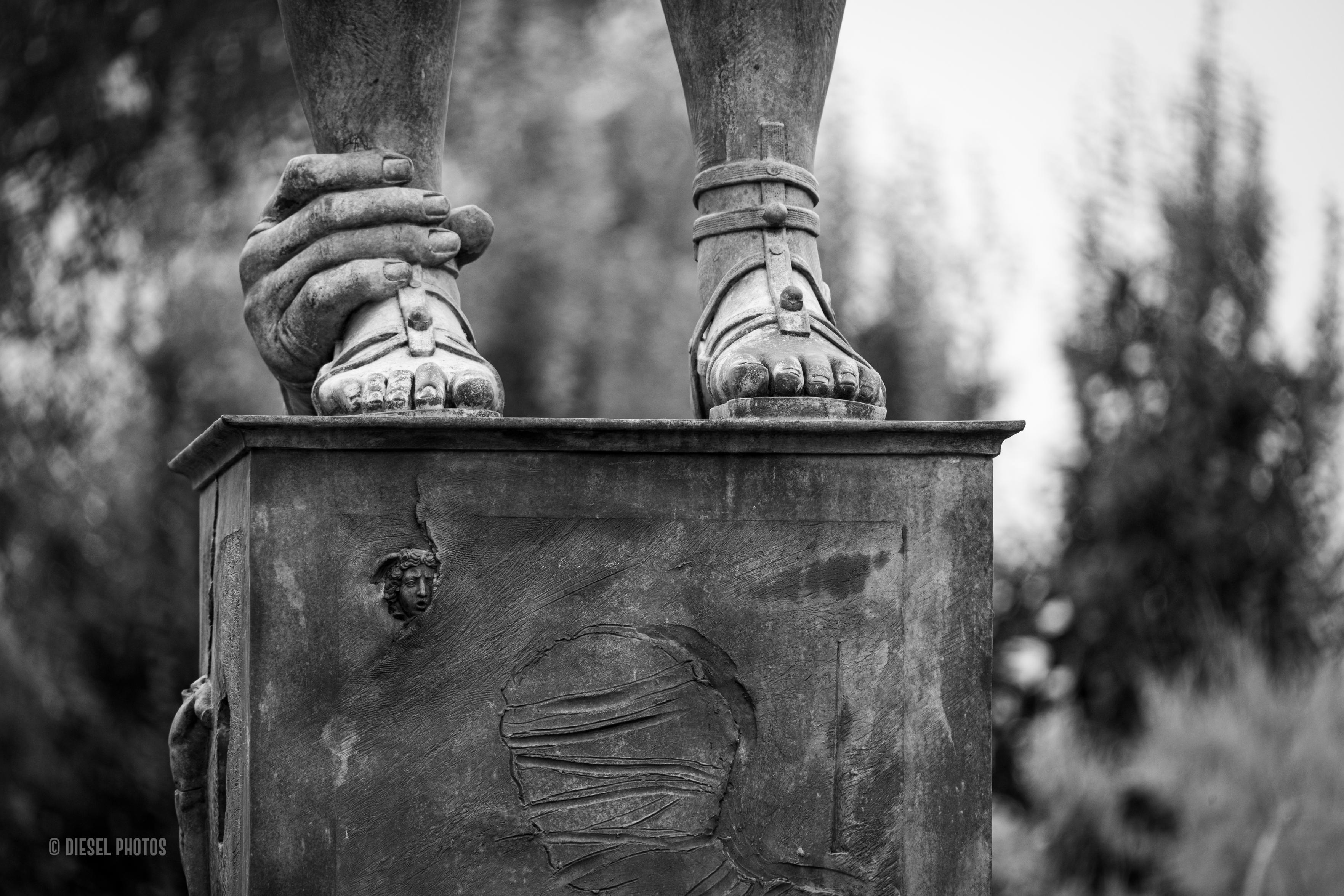 Fragment monumentalnej rzeźby ukazuje stopy odziane w antyczne sandały stojące na wysokim postumencie. Jedną z kostek obejmuje nienaturalnie duża dłoń, tworząc niepokojący, surrealistyczny kontrast między spokojną pozą a symbolicznym gestem zatrzymania. Na przedniej ścianie cokołu widoczny jest płaskorzeźbiony zarys zabandażowanej twarzy oraz niewielka klasyczna maska umieszczona wyżej. Czarno-biała fotografia wydobywa fakturę metalu i subtelne ślady patyny, a miękko rozmyte tło z drzewami kieruje uwagę wyłącznie na niezwykłą kompozycję rzeźby. Całość sprawia wrażenie zagadki pozostawionej widzowi do własnej interpretacji.