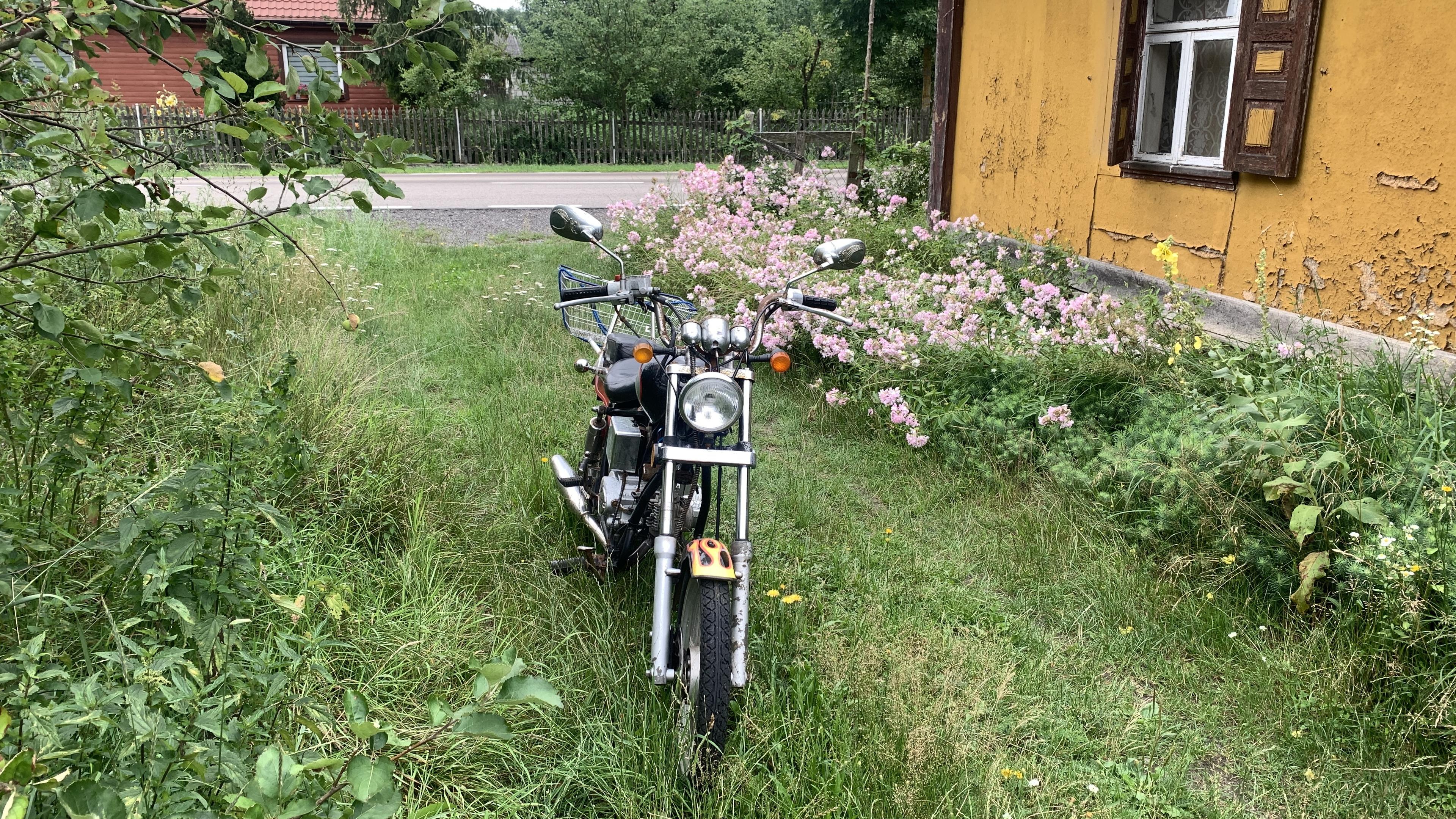 Motorek typu chopper przed starym drewnianym domem, na trawie, obok kwiatów
