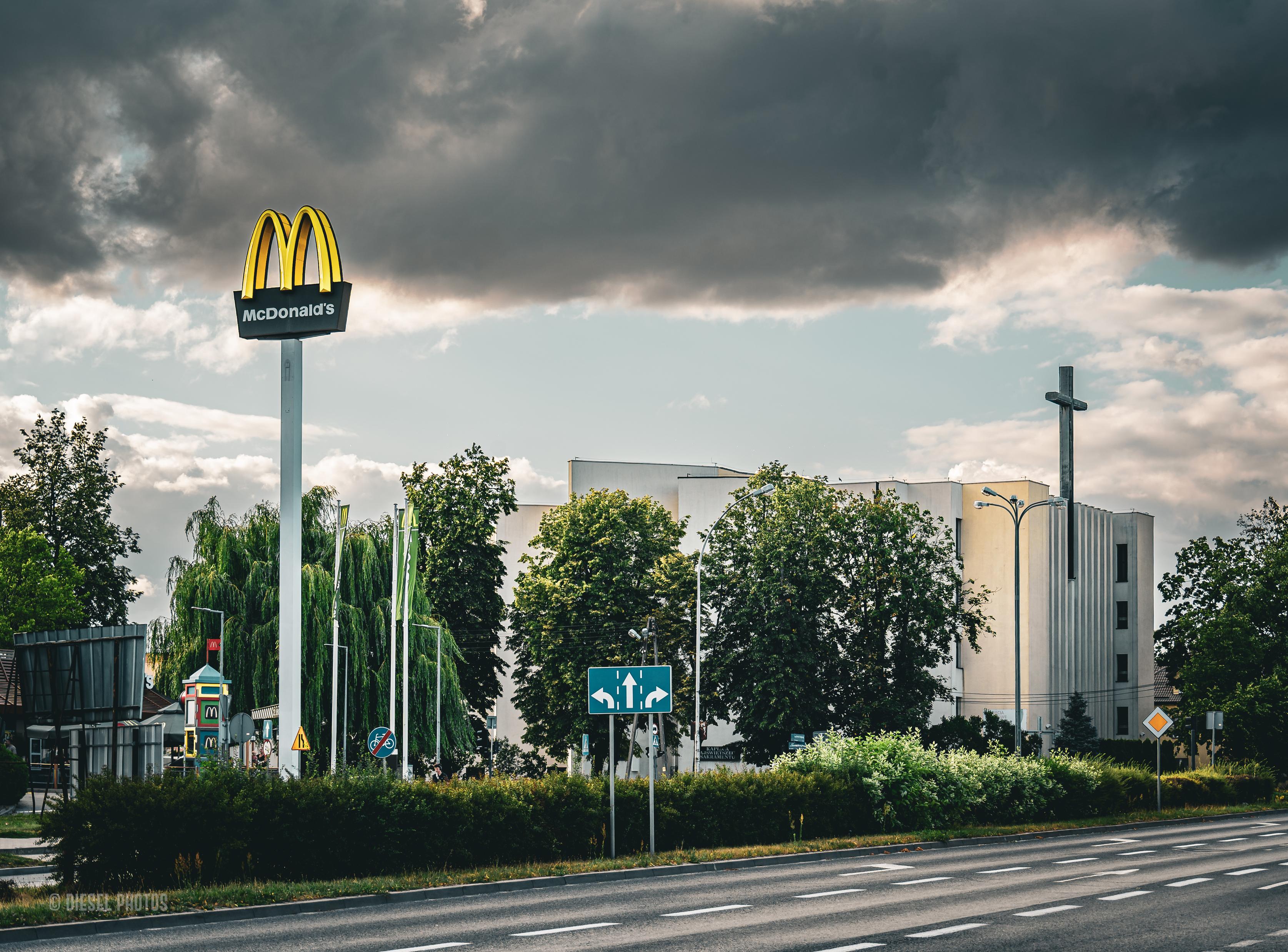 Po szerokiej, wielopasmowej ulicy nie jedzie żaden samochód. W centrum kadru, na tle jasnego budynku, stoją dwa wysokie symbole wyrastające ponad korony drzew. Po lewej stronie znajduje się charakterystyczny znak McDonald’s – złote łuki na wysokim białym słupie. Po prawej stronie wznosi się prosty, ciemny krzyż umieszczony przy nowoczesnym kościele.

Największą część górnej połowy zdjęcia zajmuje ciężka, niemal czarna chmura burzowa, która zwisa nisko nad miastem. Pod nią pozostaje wąski pas jasnego, błękitnego nieba z białymi obłokami. Zieleń drzew kontrastuje z ciemnym niebem i białą bryłą kościoła.

Kompozycja jest oszczędna i bardzo symboliczna – dwa pionowe znaki stoją po przeciwnych stronach kadru niczym dwa punkty odniesienia współczesnego krajobrazu. Ciemne chmury nadają fotografii dramatyczny, nieco niepokojący nastrój, sprawiając, że zwykły miejski widok nabiera metaforycznego znaczenia.