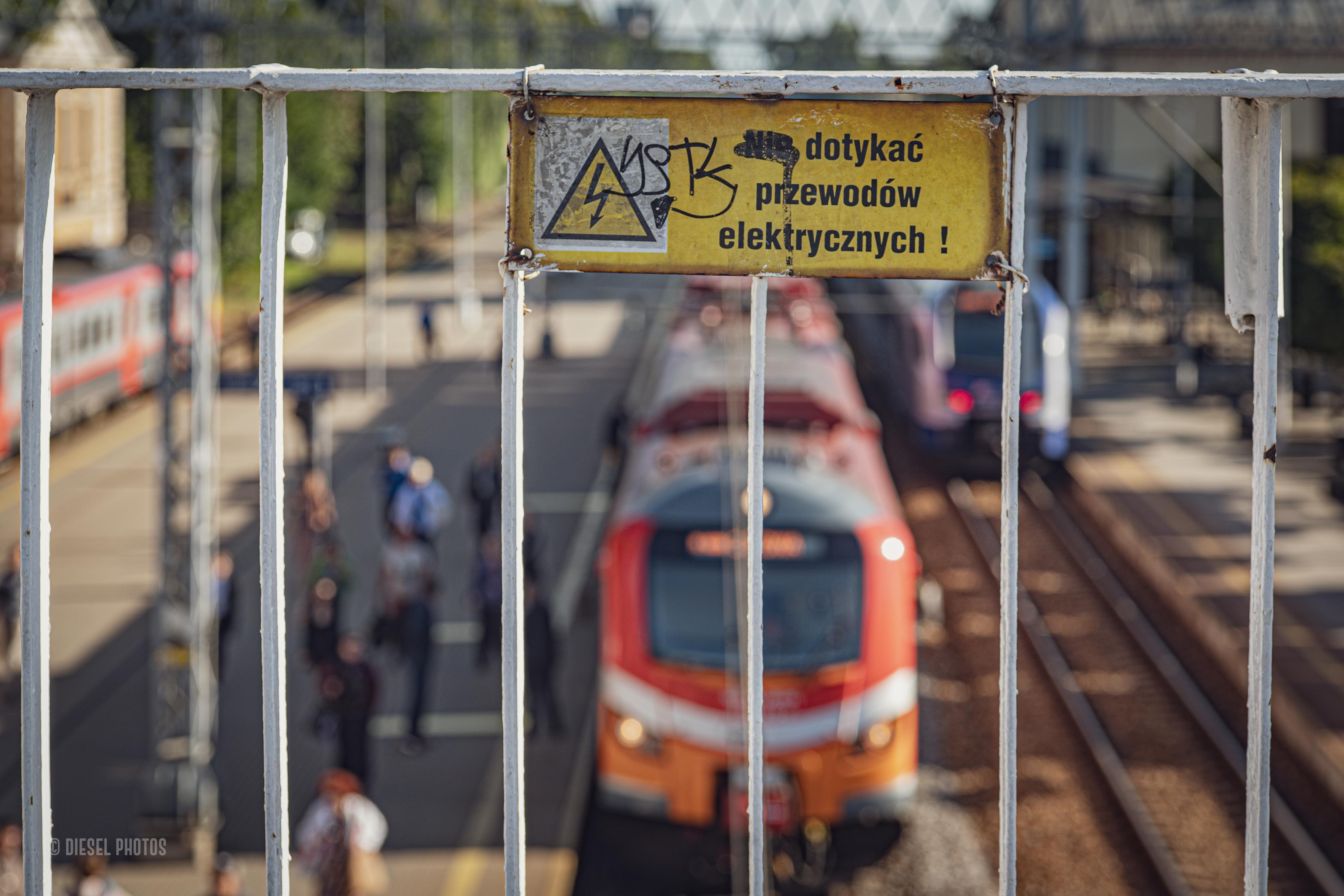 Fotografia dworca z pociągami. Wszystko jest lekko rozmyte z wyjątkiem tabliczki z informacją że przewody trakcyjne są pod napięciem.