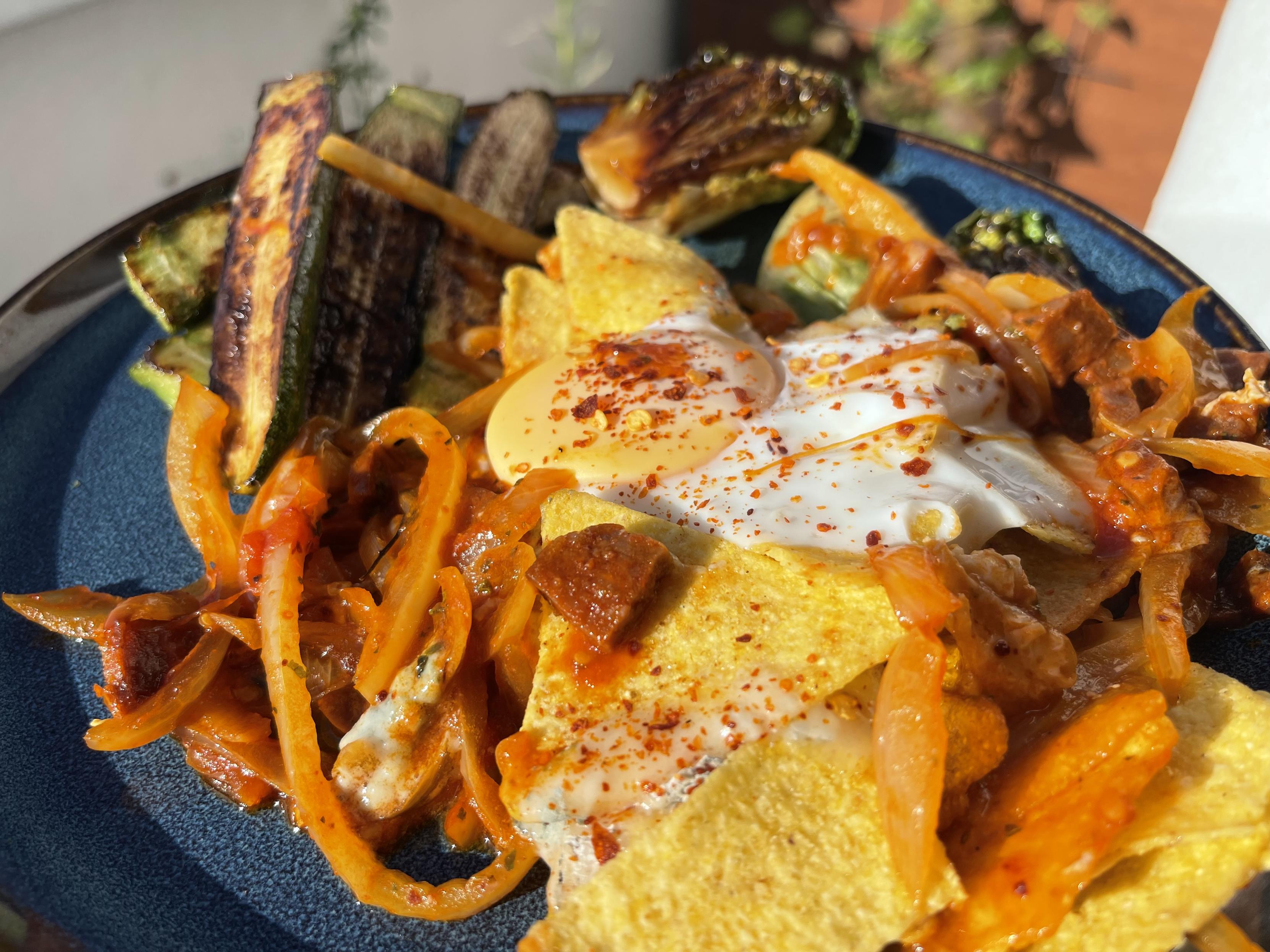 porcja chilaquiles z chrupiącymi kawałkami tortilli w czerwonym sosie z papryką ramiro i cebulą. Na wierzchu znajduje się jajko sadzone z płynnym żółtkiem posypane płatkami chili. Obok ułożono grillowane mini cukinie oraz młodą sałatę rzymską z wyraźnie przypieczonymi brzegami.