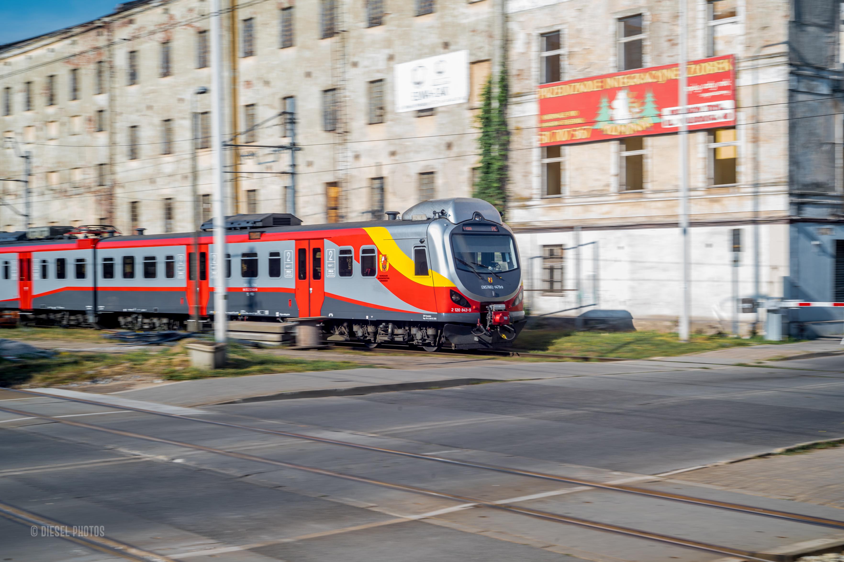 Na zdjęciu widać nowoczesny pociąg pasażerski jadący przez miasto. Dzięki zastosowaniu panoramowania skład pozostaje stosunkowo ostry, podczas gdy tło – stara, wielokondygnacyjna kamienica oraz elementy infrastruktury kolejowej – ulega charakterystycznemu rozmyciu, podkreślając wrażenie prędkości. Na pierwszym planie znajdują się tory tramwajowe i fragment jezdni, a za pociągiem widoczny jest zniszczony budynek z licznymi śladami upływu czasu. Kontrast między nowoczesnym taborem a zaniedbaną architekturą tworzy interesujące zestawienie dwóch różnych epok miejskiego krajobrazu.
