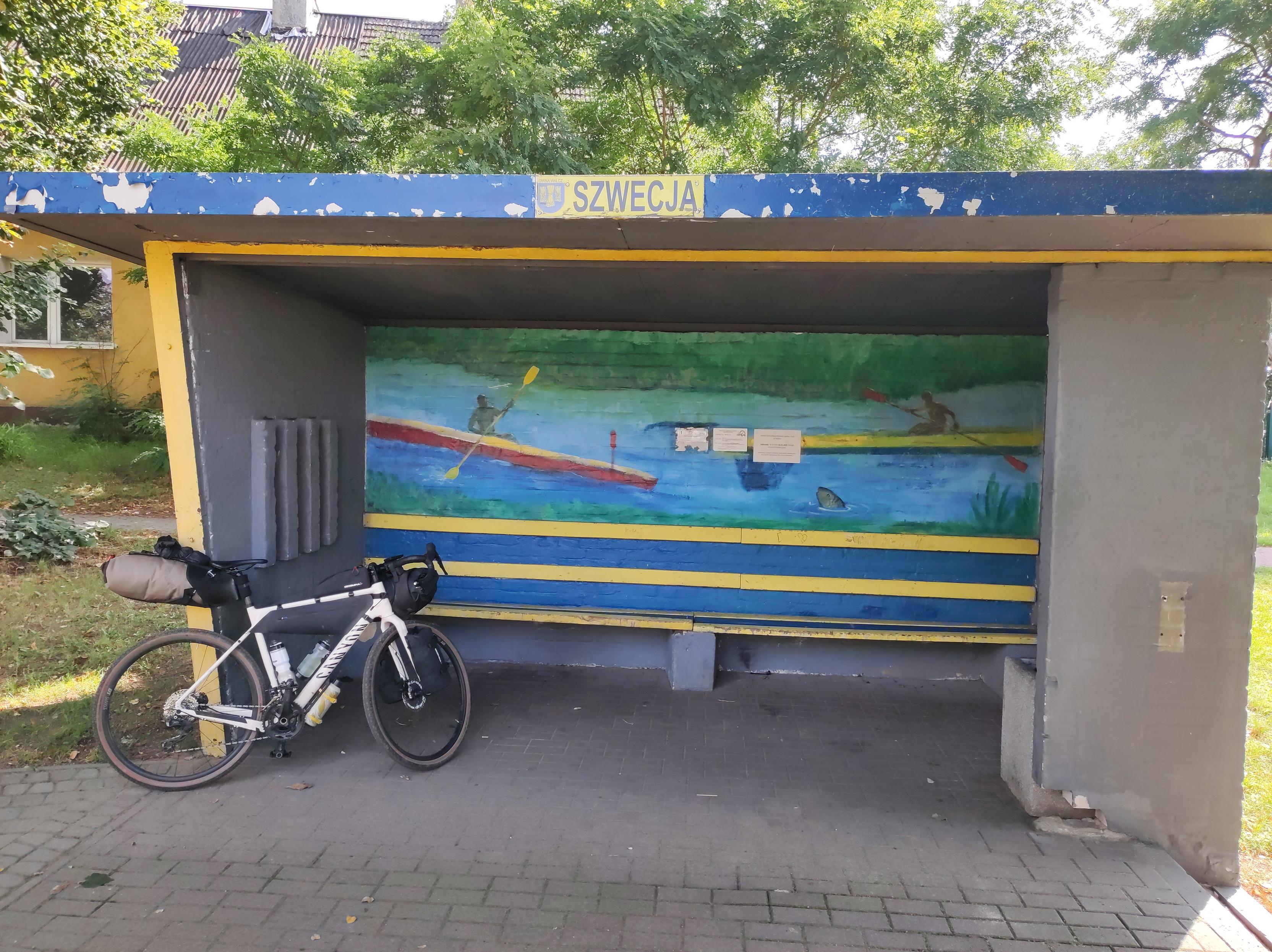 Rower oparty o wiatę przystankowa z napisem Szwecja. Wewnętrzna ściana jest pomalowana i przedstawia osobę płynącą kajakiem po rzece.