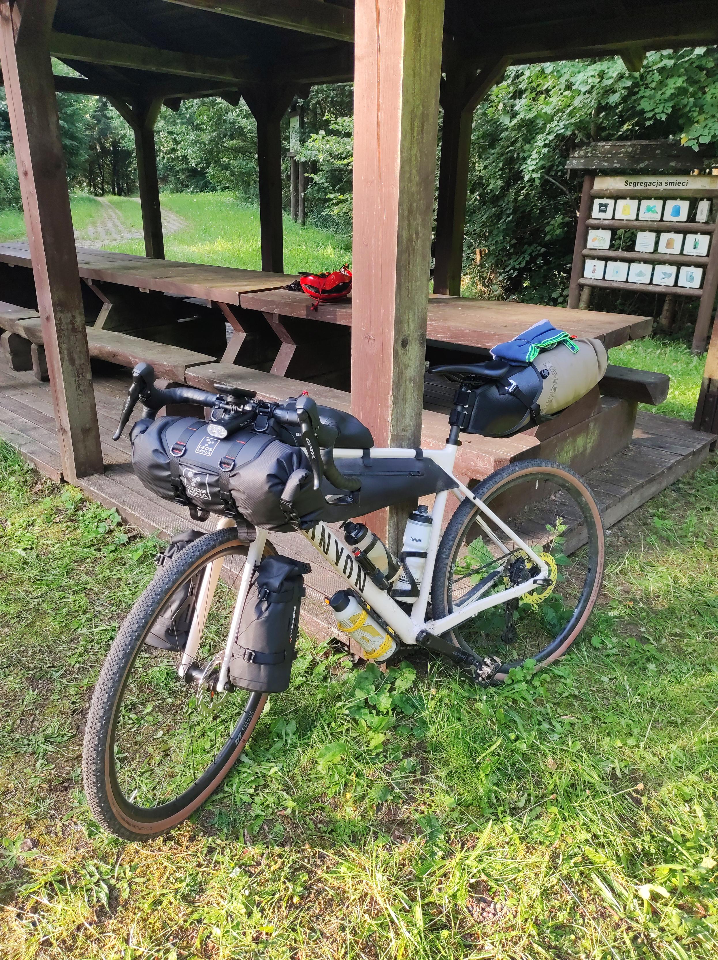 O drewnianą wiatę opiera się mój rower szutrowy Canyon, objuczony torbami. Na stole w tle leży czerwony kask kolarski.