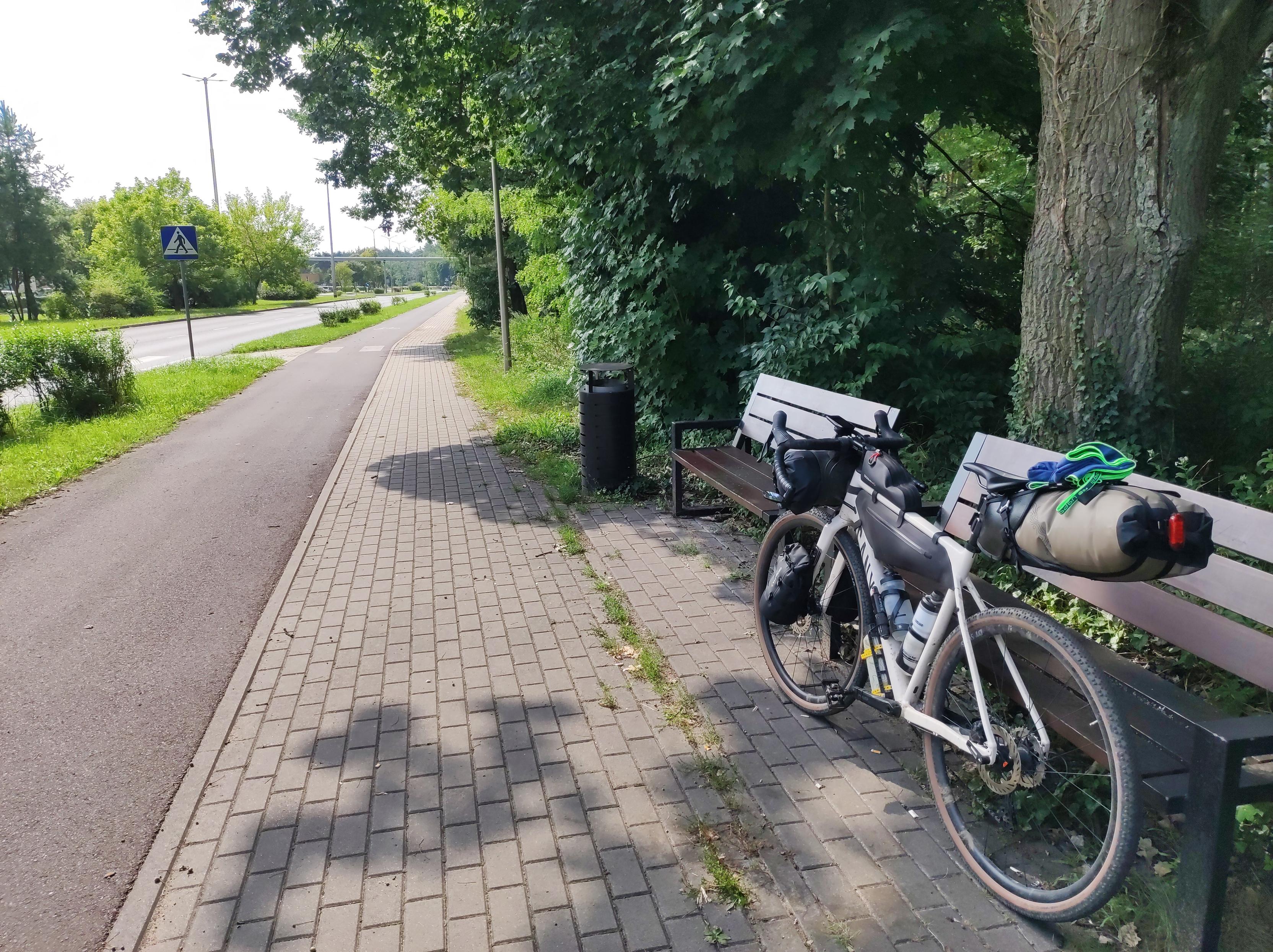 Objuczny rower szutrowy oparty o ławkę na skraju chodnika i drogi dla rowerów.