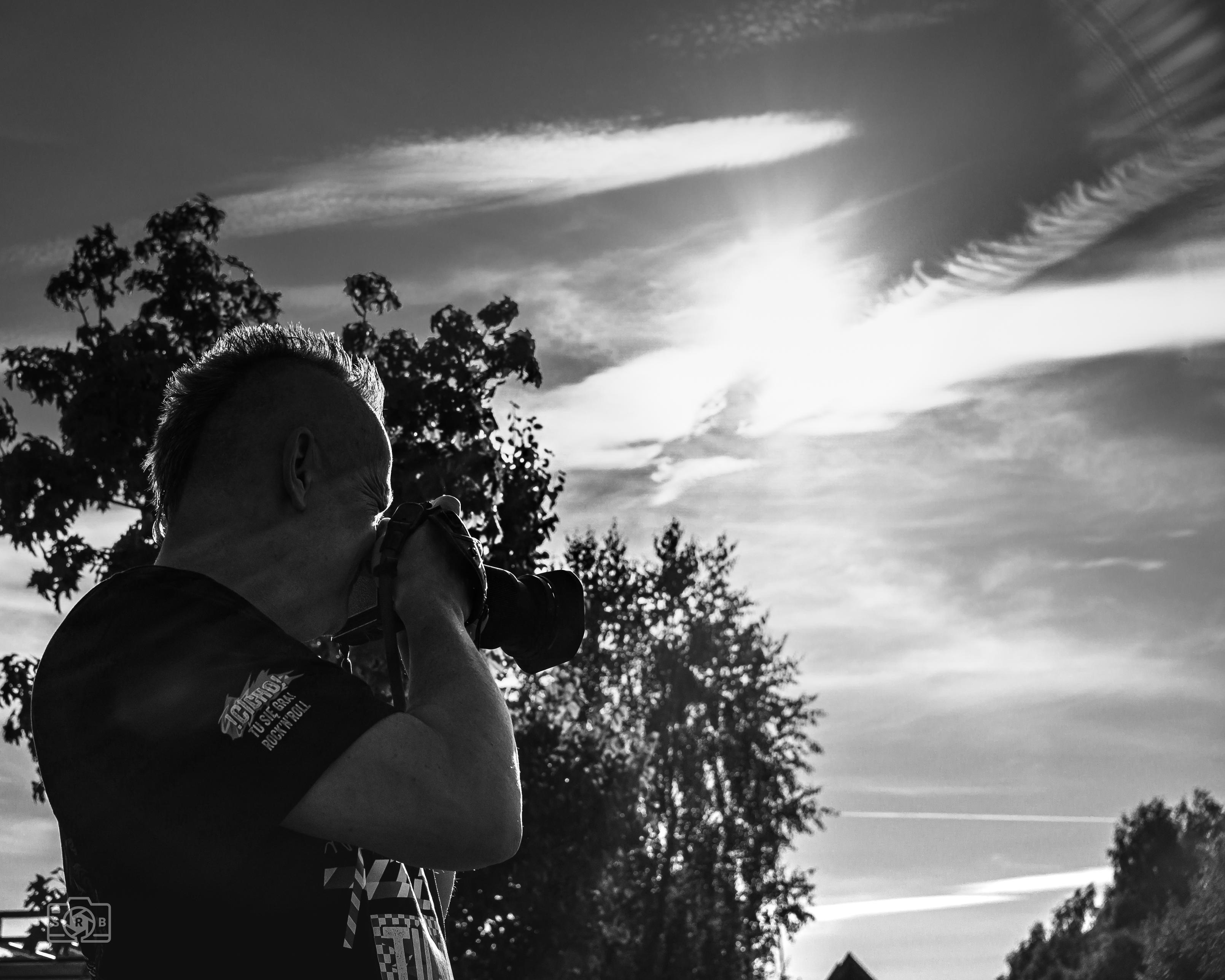 Czarno-biała fotografia przedstawia fotografa uchwyconego z profilu w chwili wykonywania zdjęcia. Sylwetka znajduje się po lewej stronie kadru, natomiast pozostałą przestrzeń wypełnia niebo z mocnym słońcem przebijającym się przez warstwy chmur i smugi kondensacyjne. Kontrast między ciemną postacią a jasnym niebem tworzy wyraźny efekt sylwetki, a drzewa stanowią naturalną ramę dla sceny.