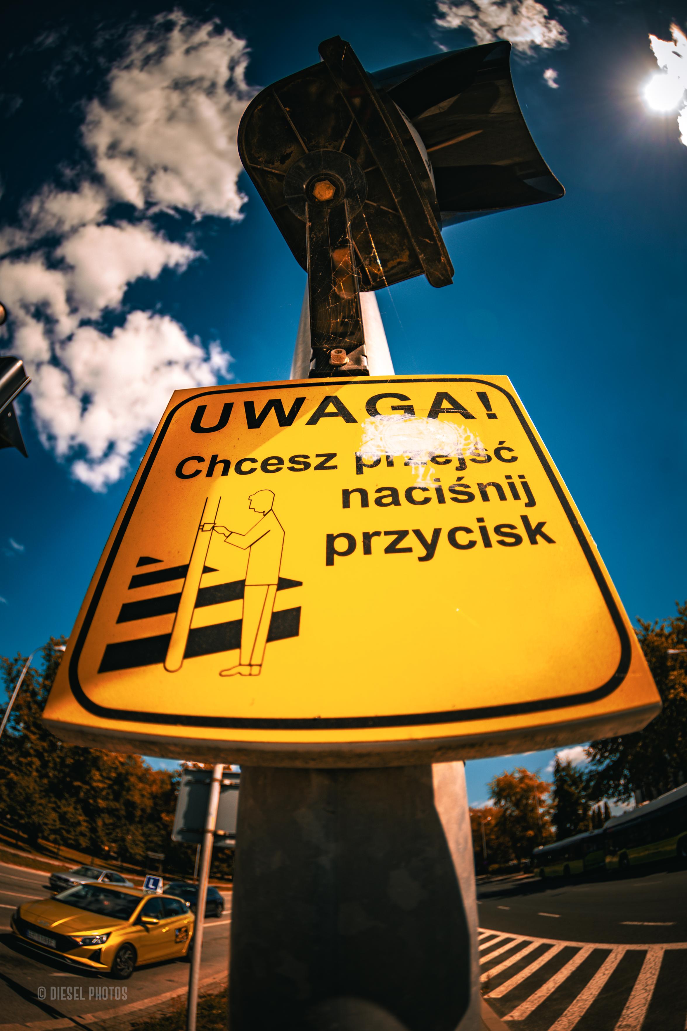 To zdjęcie przedstawia żółtą tabliczkę i instrukcją dla pieszych przy przejściu przez jezdnię sfotografowaną z bardzo niskiej perspektywy, szerokokątnym obiektywem. Tabliczka z napisem „UWAGA! Chcesz przejść – naciśnij przycisk” dominuje w kadrze, a nad nią znajduje się sygnalizator świetlny. W tle widać intensywnie niebieskie niebo z białymi chmurami oraz fragmenty ulicy z samochodami i autobusem. Dzięki mocnemu kontrastowi żółci znaku z błękitem nieba i nietypowej perspektywie zwykły element infrastruktury miejskiej staje się głównym bohaterem fotografii.