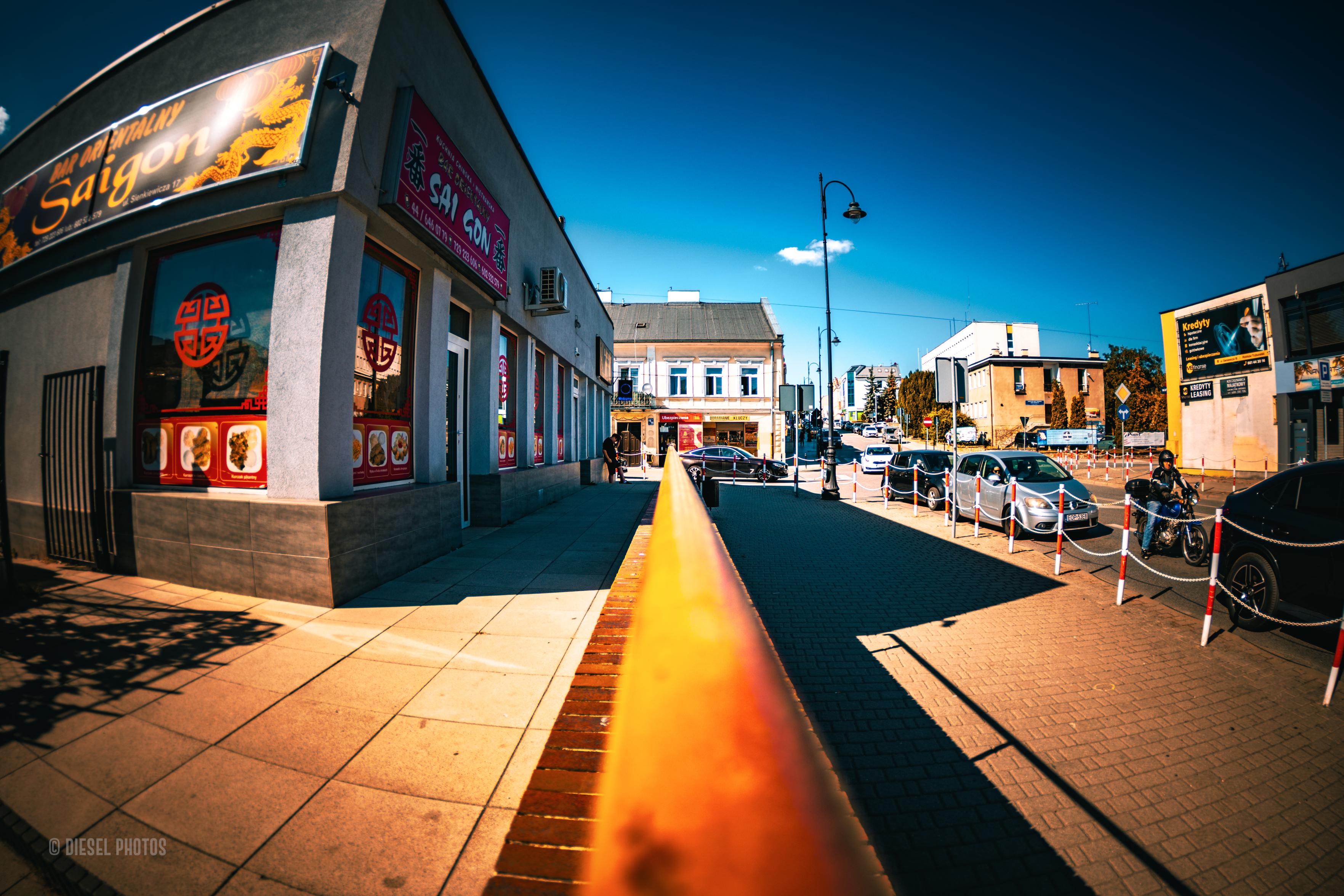 To zdjęcie przedstawia fragment miejskiej ulicy sfotografowany bardzo szerokokątnym obiektywem z perspektywy prowadnicy lub poręczy biegnącej przez środek kadru. Żółto-pomarańczowa poręcz tworzy mocną linię prowadzącą wzrok w głąb ulicy. Po lewej stronie znajduje się restauracja orientalna z dużymi witrynami i kolorowymi szyldami, po prawej zaparkowane samochody oraz latarnie uliczne. W tle widać niską zabudowę i intensywnie niebieskie niebo z pojedynczą białą chmurą. Charakterystyczne zniekształcenia szerokokątnego obiektywu podkreślają dynamikę kompozycji, sprawiając, że zwykła miejska scena nabiera geometrycznego i niemal futurystycznego charakteru.