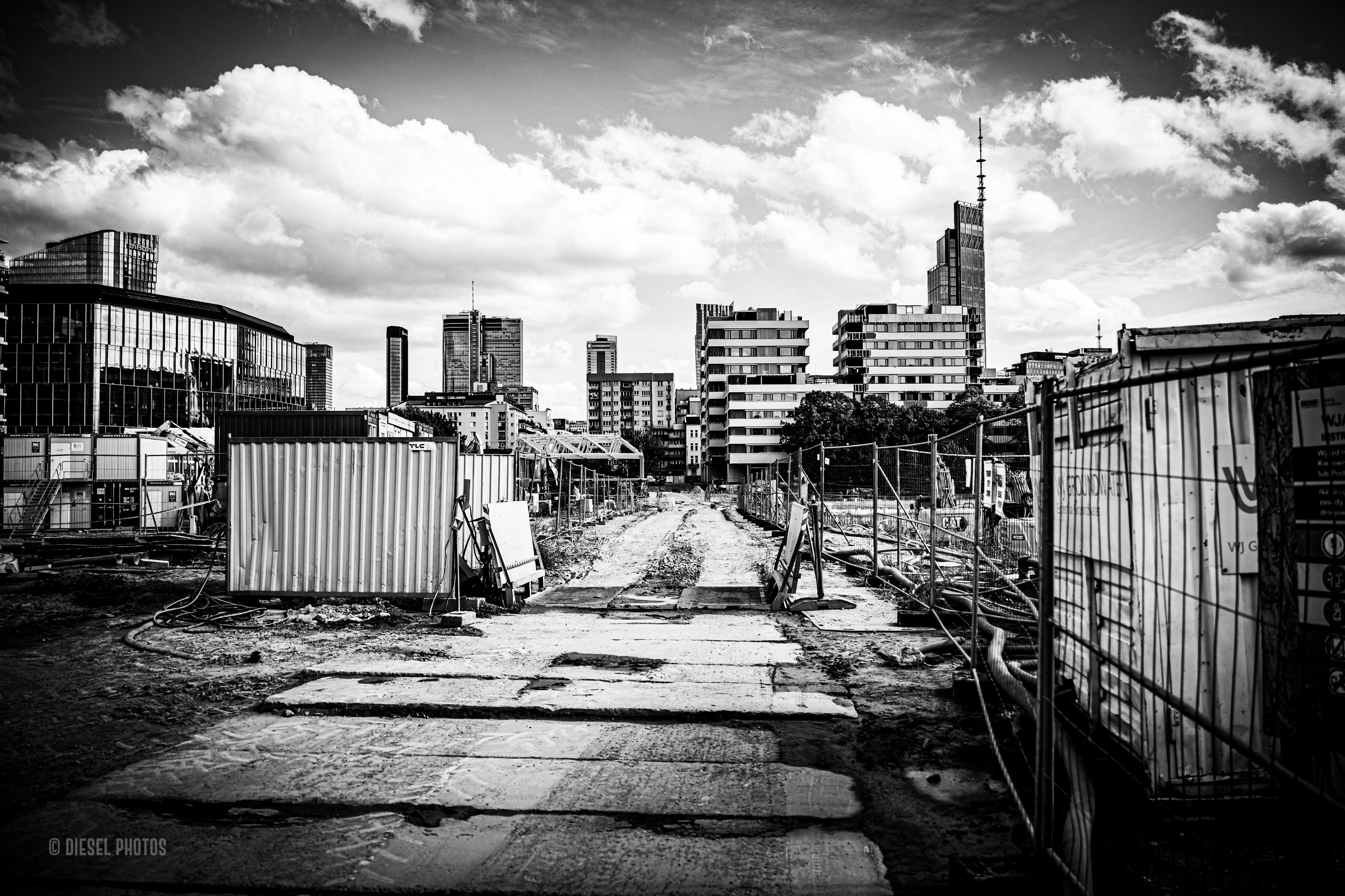 Czarno-białe, poziome zdjęcie przedstawia rozległy plac budowy w centrum Warszawy. Fotograf stoi na początku prowizorycznej drogi z dużych, nierównych betonowych płyt. Droga biegnie przez środek kadru i zwęża się wraz z perspektywą, prowadząc wzrok wprost ku panoramie miasta.
Po obu stronach drogi znajduje się zaplecze budowy: metalowe ogrodzenia, kontenery, przewody, rury, fragmenty betonu i rozkopany teren. Wszystko jest surowe, nieuporządkowane i wyraźnie tymczasowe. Po prawej stronie duży kontener oraz ogrodzenie tworzą ciemną ścianę kadru, po lewej stoją mniejsze kontenery i elementy infrastruktury budowlanej.
W głębi, za placem budowy, wyrasta gęsta zabudowa centrum Warszawy — bloki, biurowce i wysokie wieżowce o różnych kształtach. Nowoczesna panorama miasta mocno kontrastuje z chaotycznym, brudnym placem budowy na pierwszym planie.
Górną część fotografii zajmuje bardzo wyraziste niebo z ogromnymi, jasnymi chmurami na ciemniejszym tle. Zdjęcie ma wysoki kontrast: jasne chmury, b