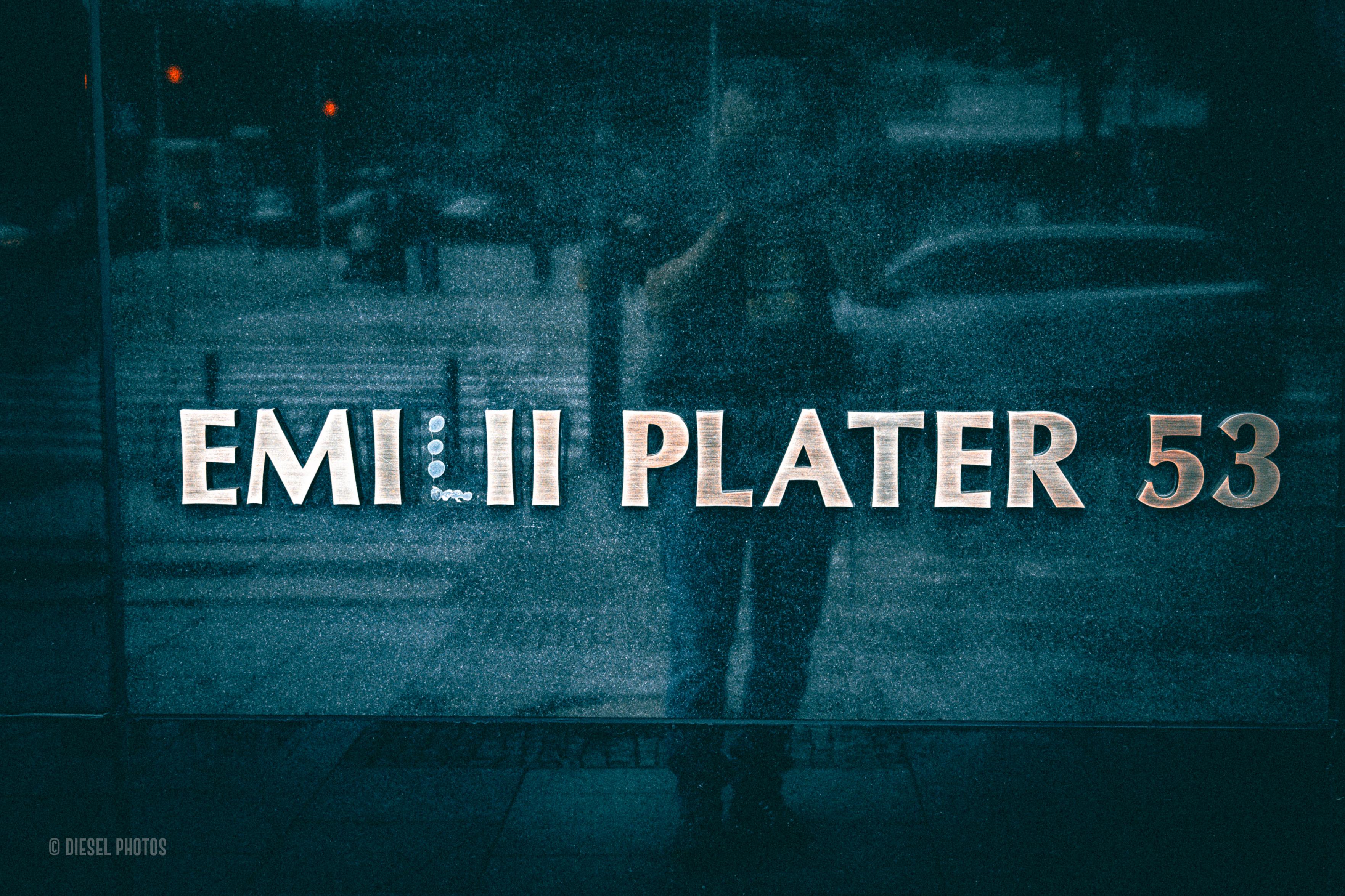 Ciemna, mocno ziarnista fotografia przedstawia szybę lub witrynę z dużym napisem „EMILII PLATER 53”. W szkle odbija się miejska ulica: samochody, fragmenty zabudowy oraz sylwetka osoby stojącej niemal dokładnie pośrodku kadru. Dominują głębokie turkusowo-niebieskie tony, przełamane jasnymi literami napisu i kilkoma drobnymi czerwonymi punktami światła w tle. Odbicia, ziarno i przygaszone kolory nadają fotografii surowy, nieco filmowy charakter.